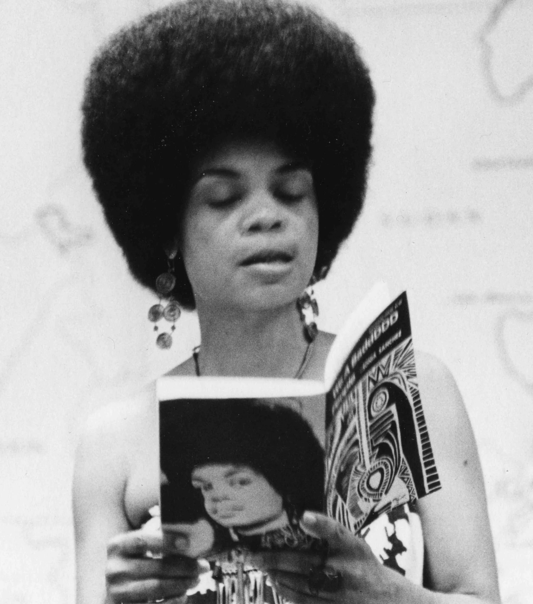 Poet Sonia Sanchez in a black and white portrait, wearing large decorative earrings and looking down at a book she is holding.