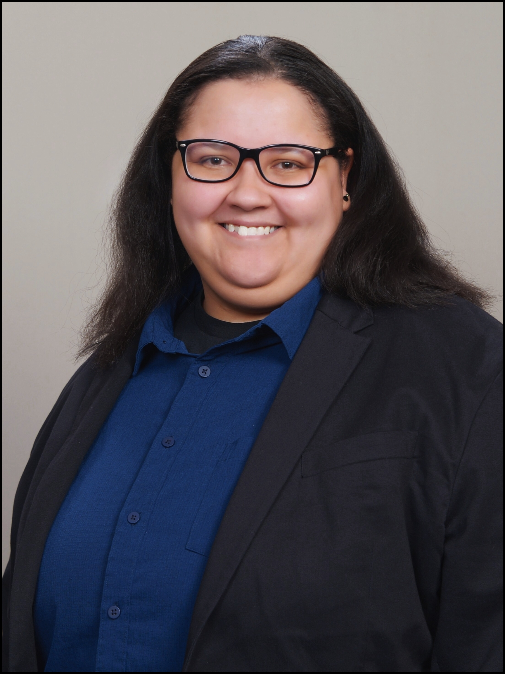 Holly E. Burgess, with long dark hair, glasses, and a dark blazer over a blue shirt, smiling in front of a plain light background.