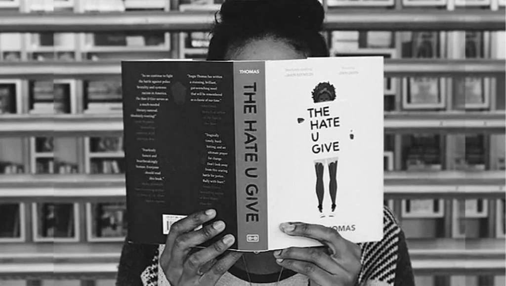 The cover of 'The Hate U Give' being read in a library setting, symbolizing the intersection of contemporary YA literature and social justice activism.