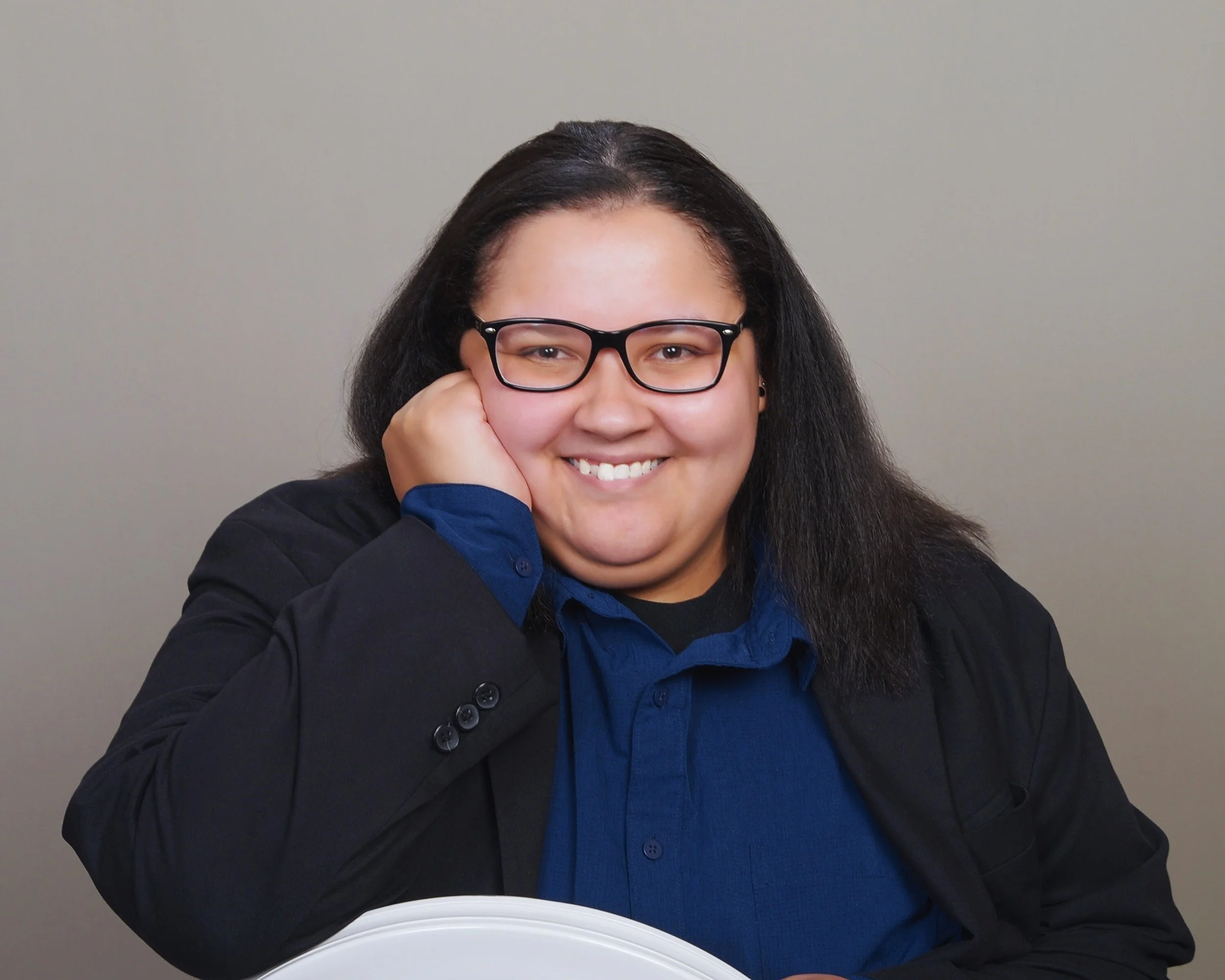 Photo of Holly Burgess seated with her hand cupping her cheek. She is stressed in a blue shirt and black blazer. She is wearing Black Raybans.