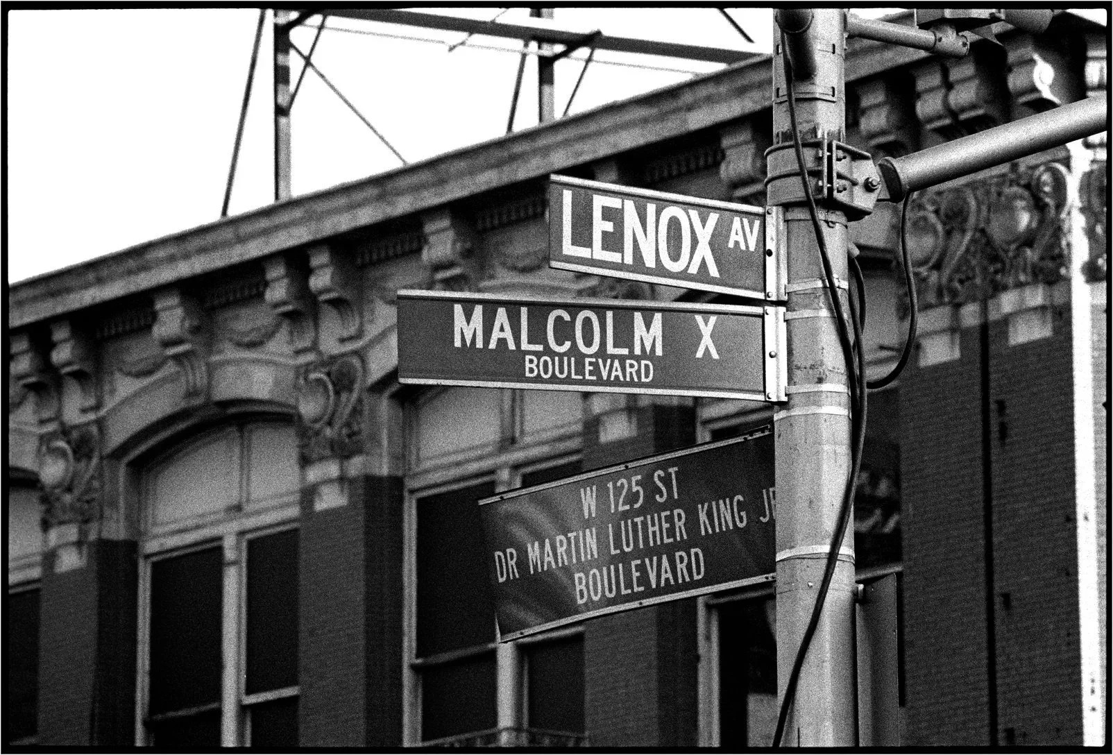 Street signs at the intersection of Lenox Avenue, Malcolm X Boulevard, and W 125th Street, with a building in the background with decorative architectural details.
