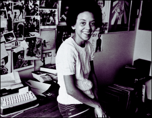 Nikki Giovanni smiles in her workspace, surrounded by a typewriter, books, and a wall of photographs.