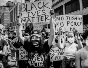 Crowd of protesters on a city street in black and white. The central figure is a woman holding a large cardboard sign that says 'BLACK LIVES MATTER.' Other protesters in masks hold similar signs against a backdrop of city buildings.