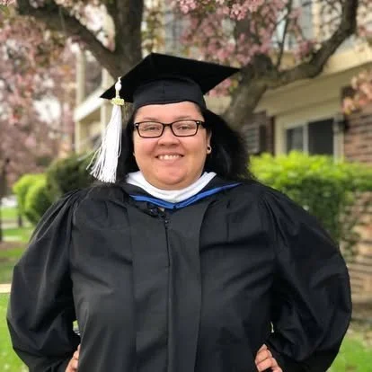 Holly E. Burgess in a cap and gown, smiling outdoors with cherry blossoms and a house in the background.