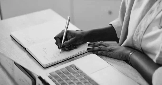 Black female's hand writing in journal with laptop next to her on a table. She is wearing a button down shirt but you only see her hands.
