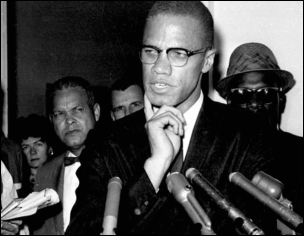 Malcolm X in a suit and glasses, gesturing toward his chin in thought, stands behind several vintage microphones while members of the press look on in a crowded room.
