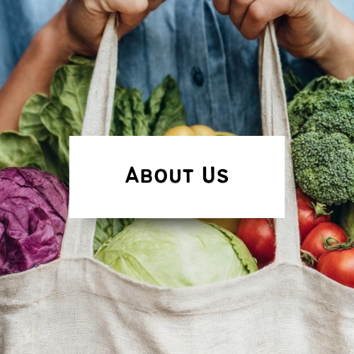 Person holding a reusable tote bag filled with fresh vegetables, including lettuce, cabbage, broccoli, and tomatoes, with a sign reading 'About Us' in front.