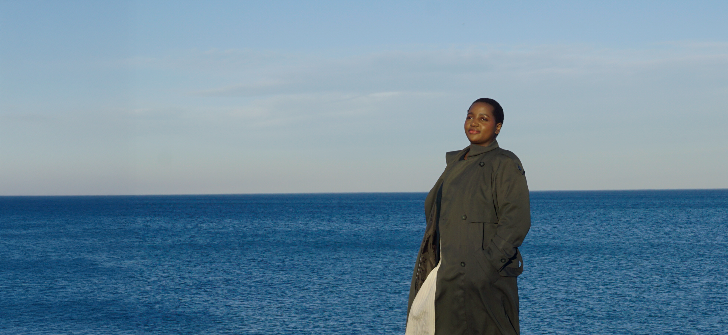 A Black woman with short hair stands on an ocean pier, wearing a long green coat, with her hands in her coat pockets, looking confidently towards the camera with a slight smile, with a clear sky and blue water visible in the background.