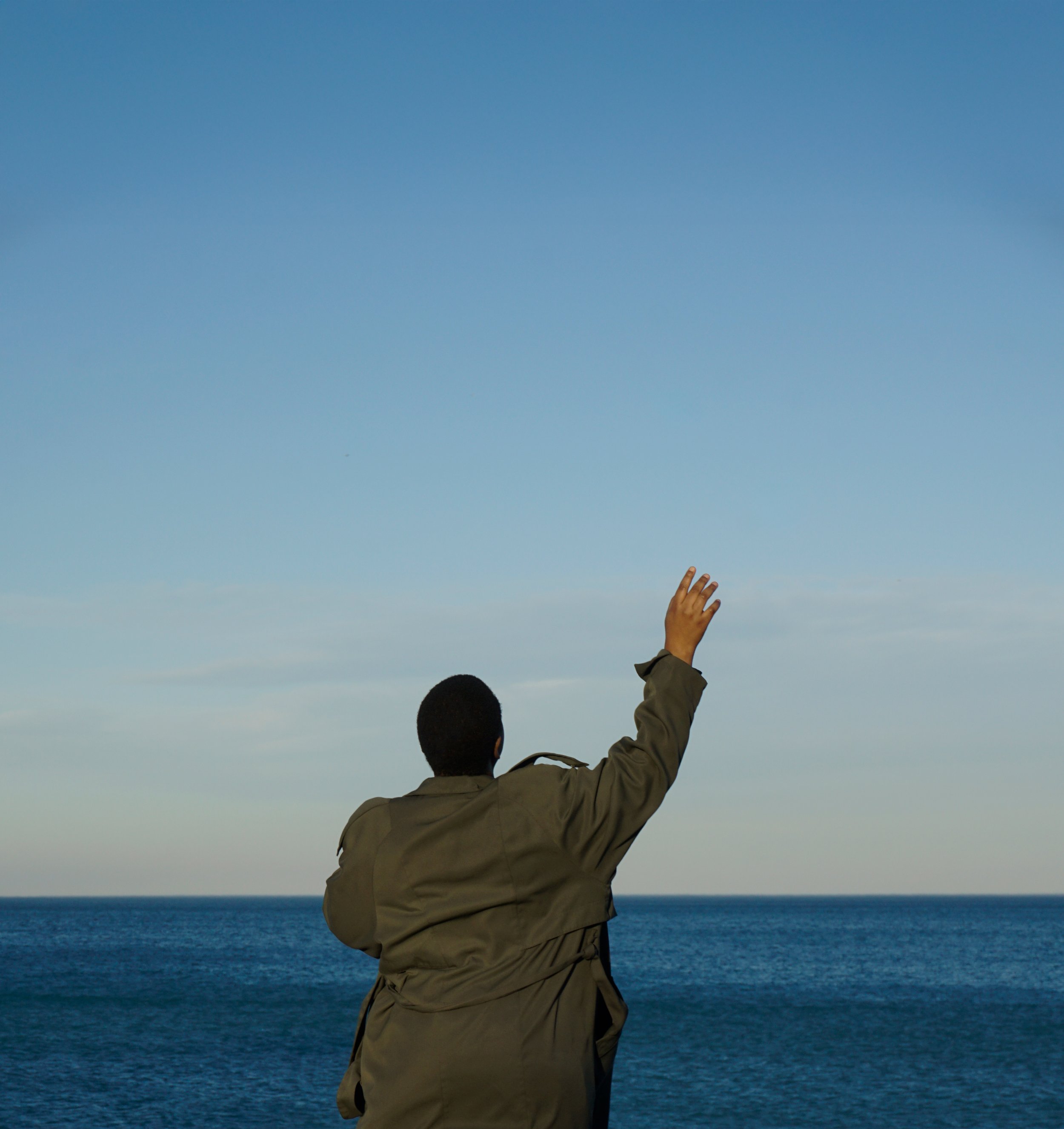 Person with short dark hair wearing a green jacket standing by the ocean, waving at the sky with their right hand on a clear day.