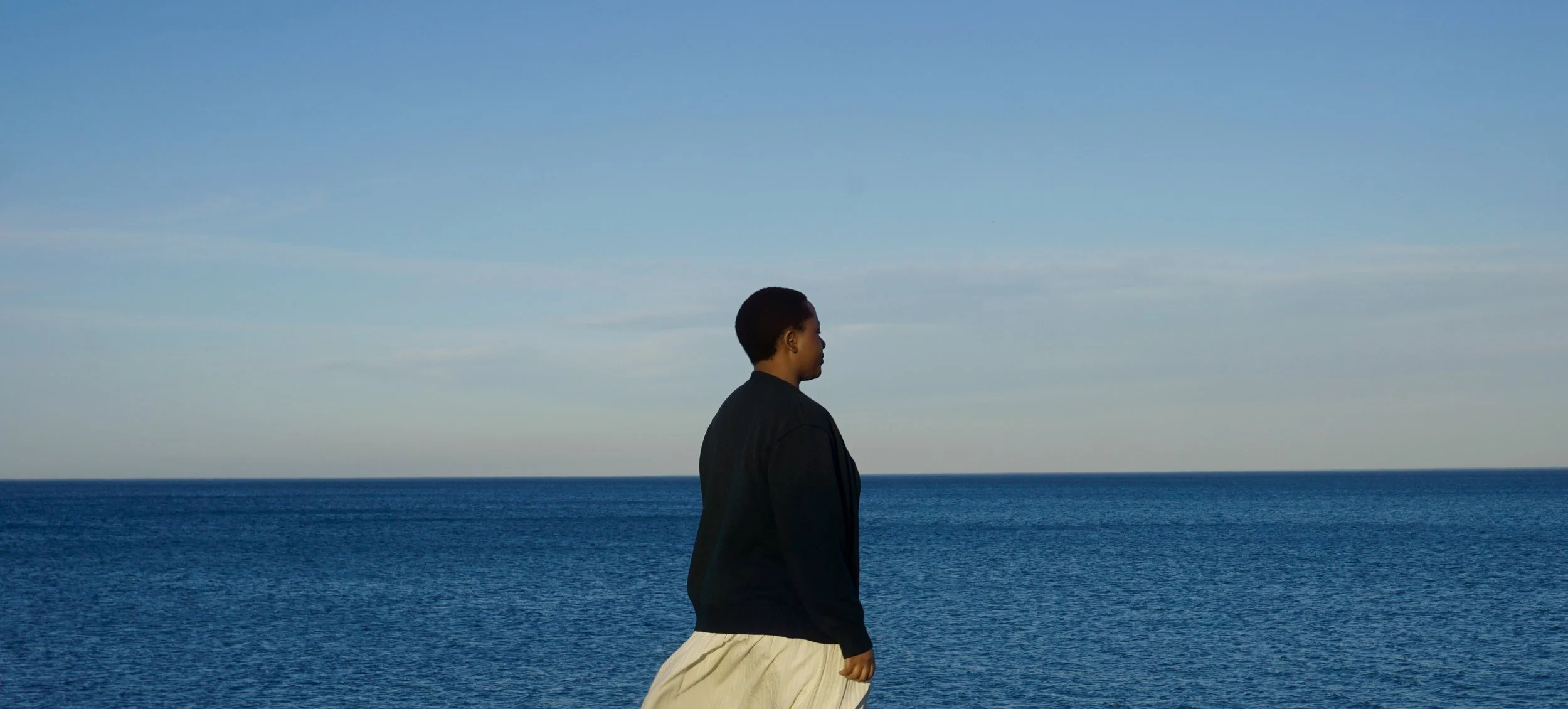 A Black woman with short hair in a black jacket and beige skirt standing by the ocean, looking towards the horizon, with a clear blue sky and blue water visible in the background.