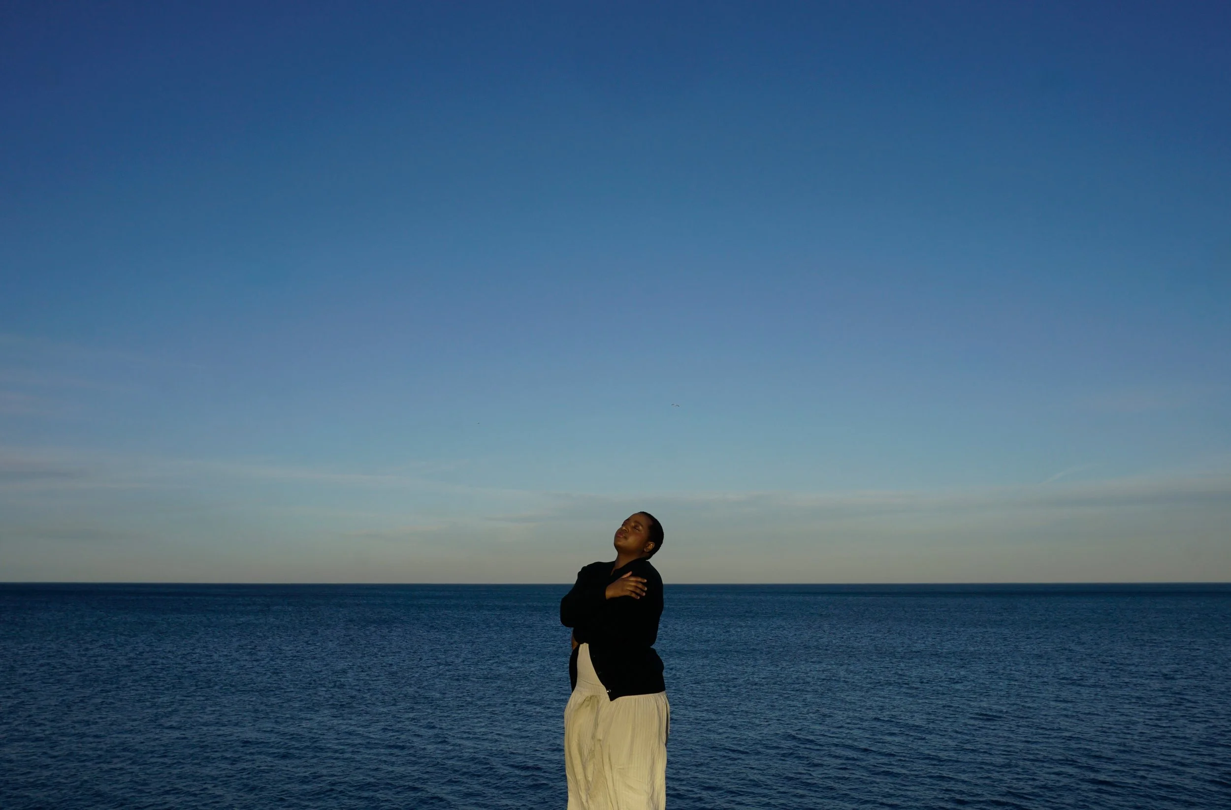 A Black woman with short hair standing by the ocean during sunset with arms crossed over her chest, gazing upward.