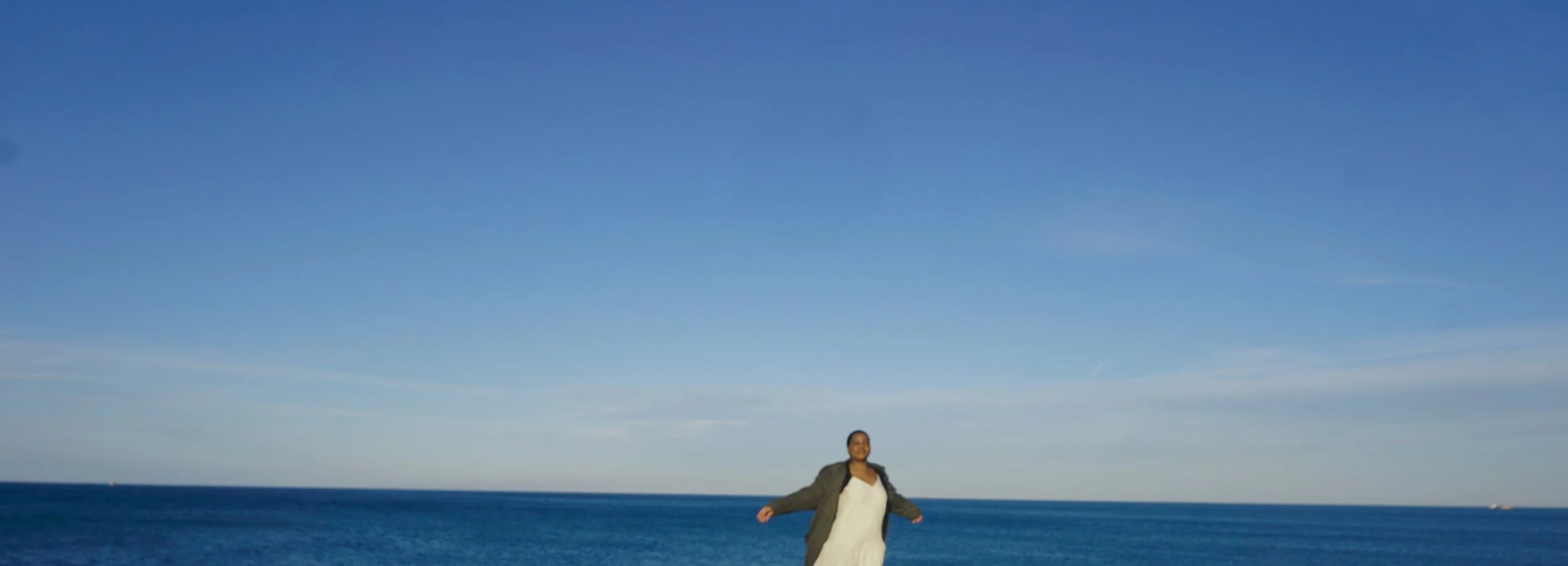 A Black woman standing on the beach with arms outstretched, ocean and blue sky in the background.