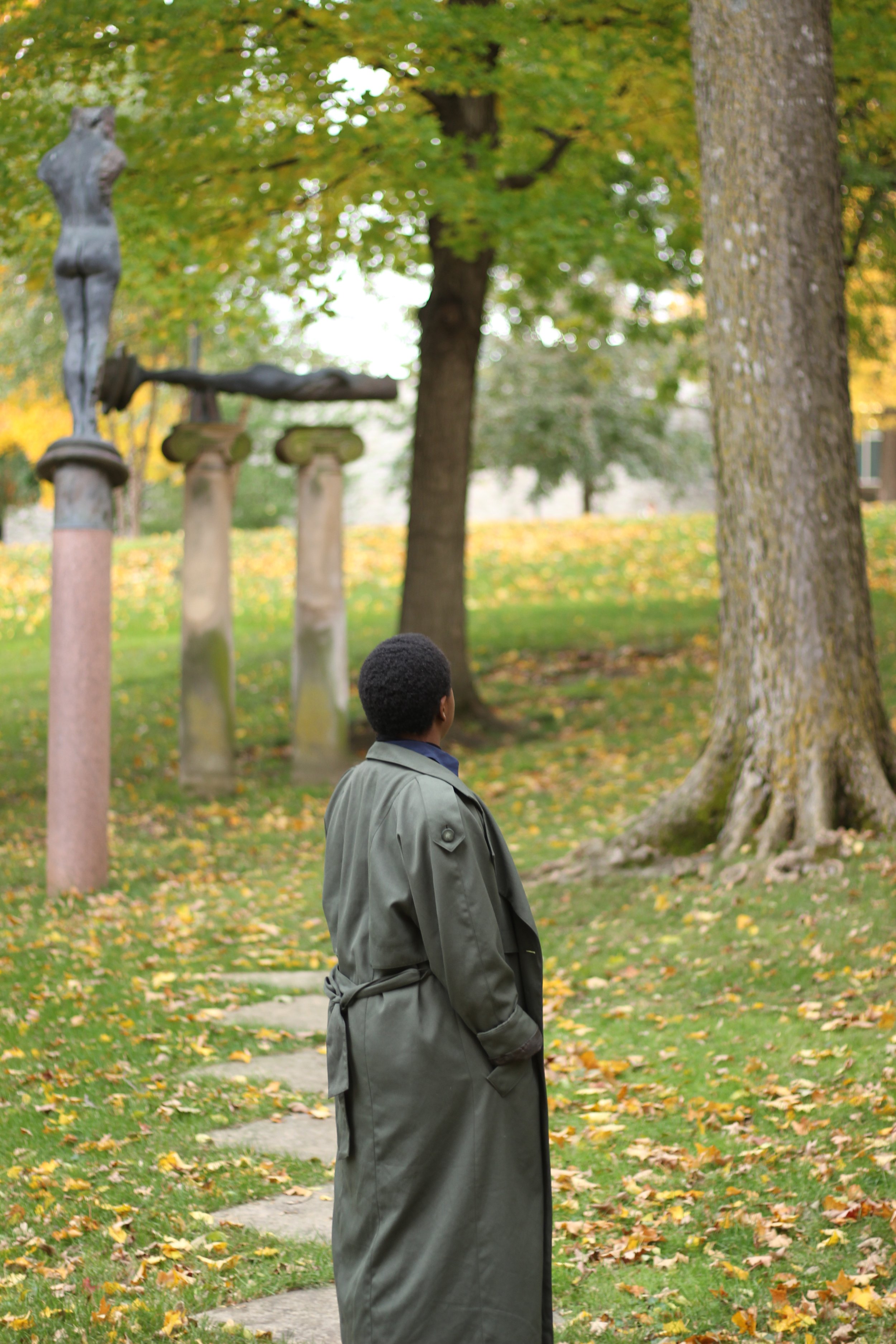 A person with short curly hair wearing a long green coat stands on a stone path in a park during fall, looking towards a statue of a woman on the left side of the image. There are trees with green and yellow leaves and fallen leaves on the ground.