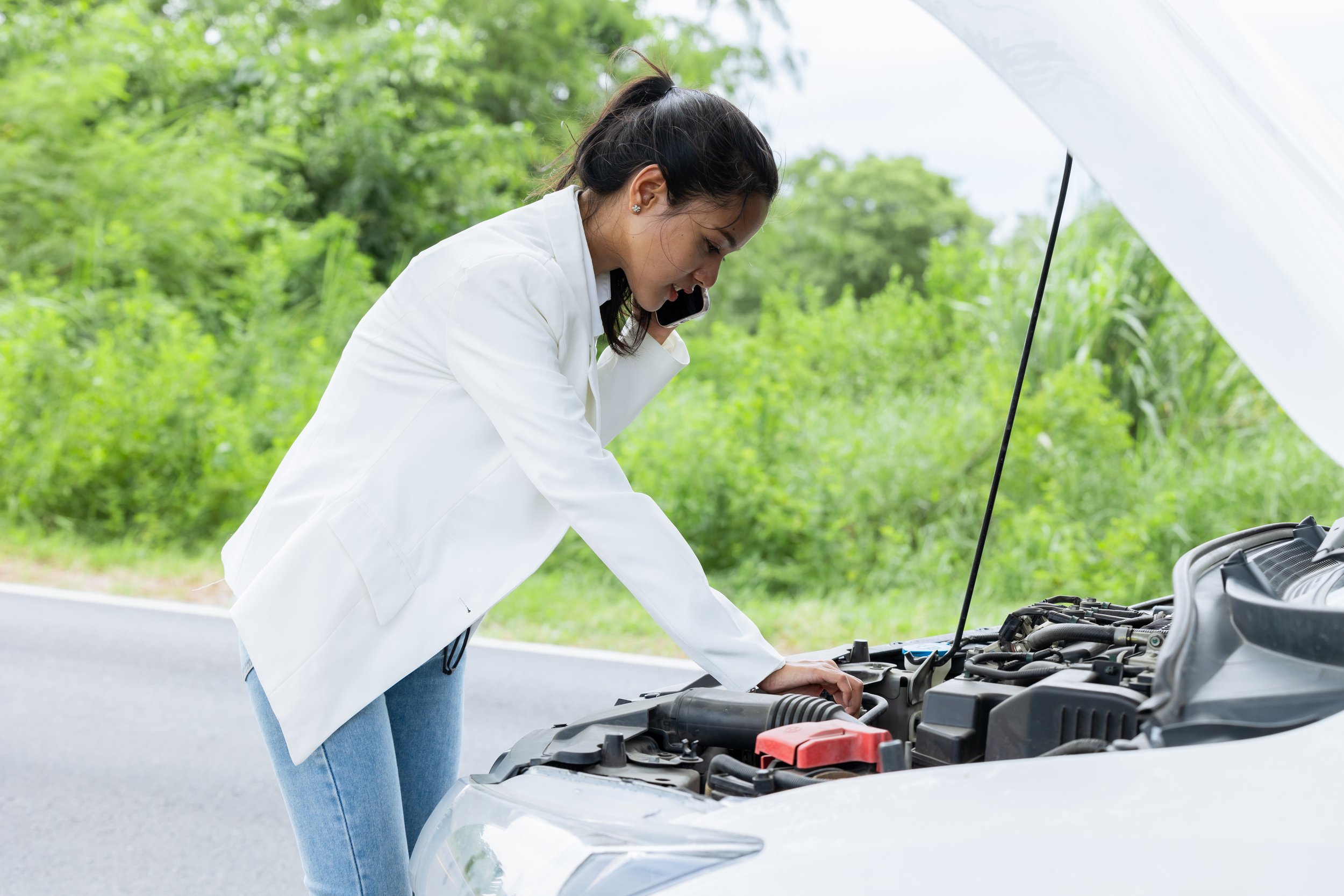 A young woman is talking on her cell phone as she stands in front of her car with the hood up , as she is broken down on the side of the road