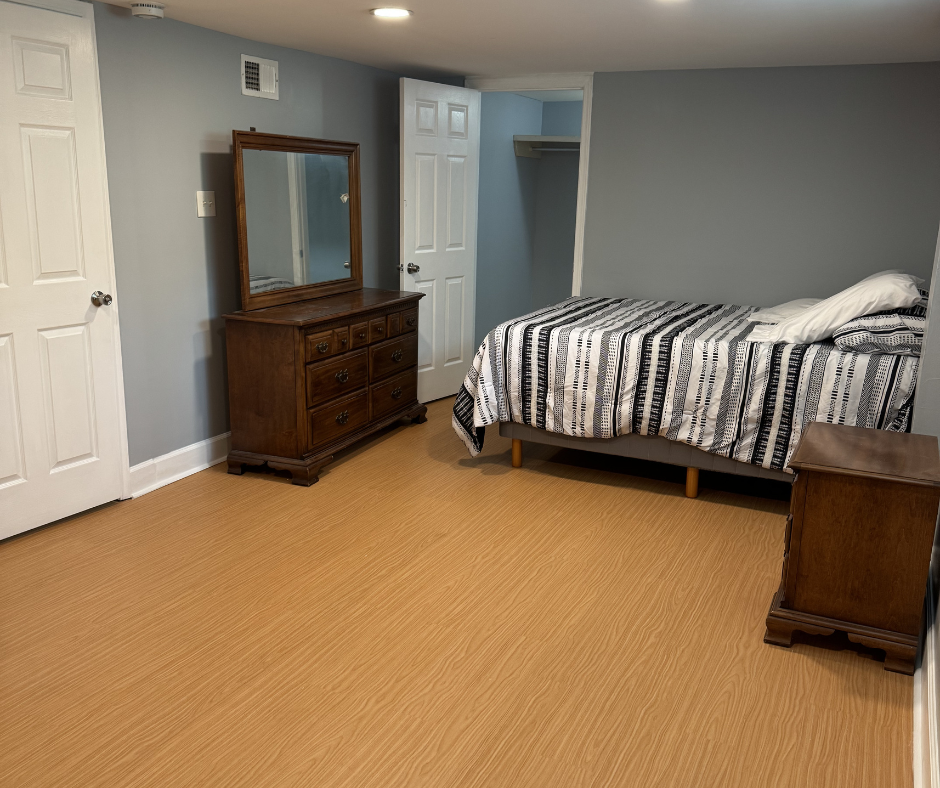 A bedroom with a bed, two wooden nightstands, a dresser with a mirror, and an open closet with a shelf, all with light blue walls and a wood laminate floor.