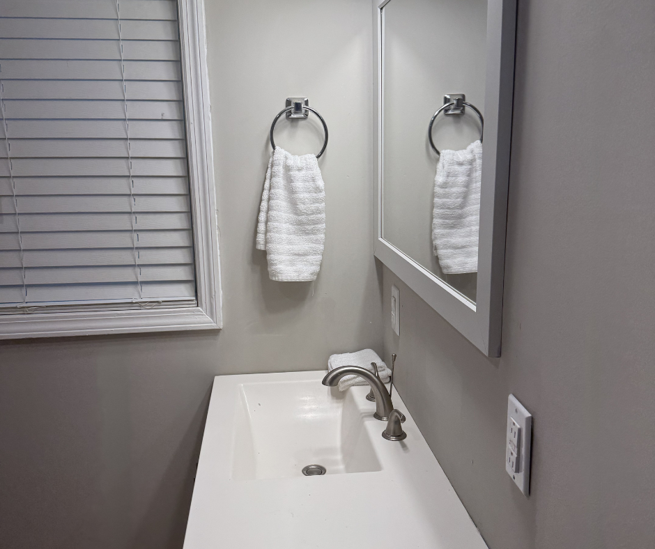 Bathroom sink with a mirror, a towel ring with a white towel, and a window with closed blinds.
