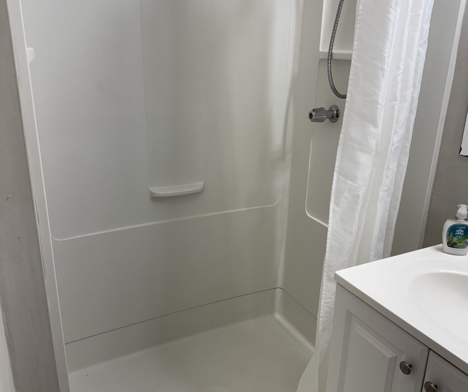 Empty shower with a white curtain, soap dish, and a handheld showerhead, adjacent to a white bathroom sink with a bottle of soap.
