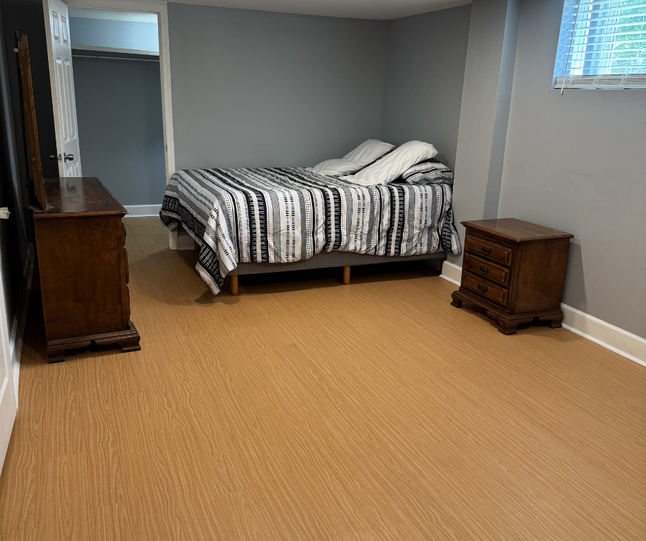 A bedroom with gray walls, a bed with a black and white striped quilt, and two wooden nightstands, one on each side of the bed. There is a small open closet on the left and a window with blinds on the right.