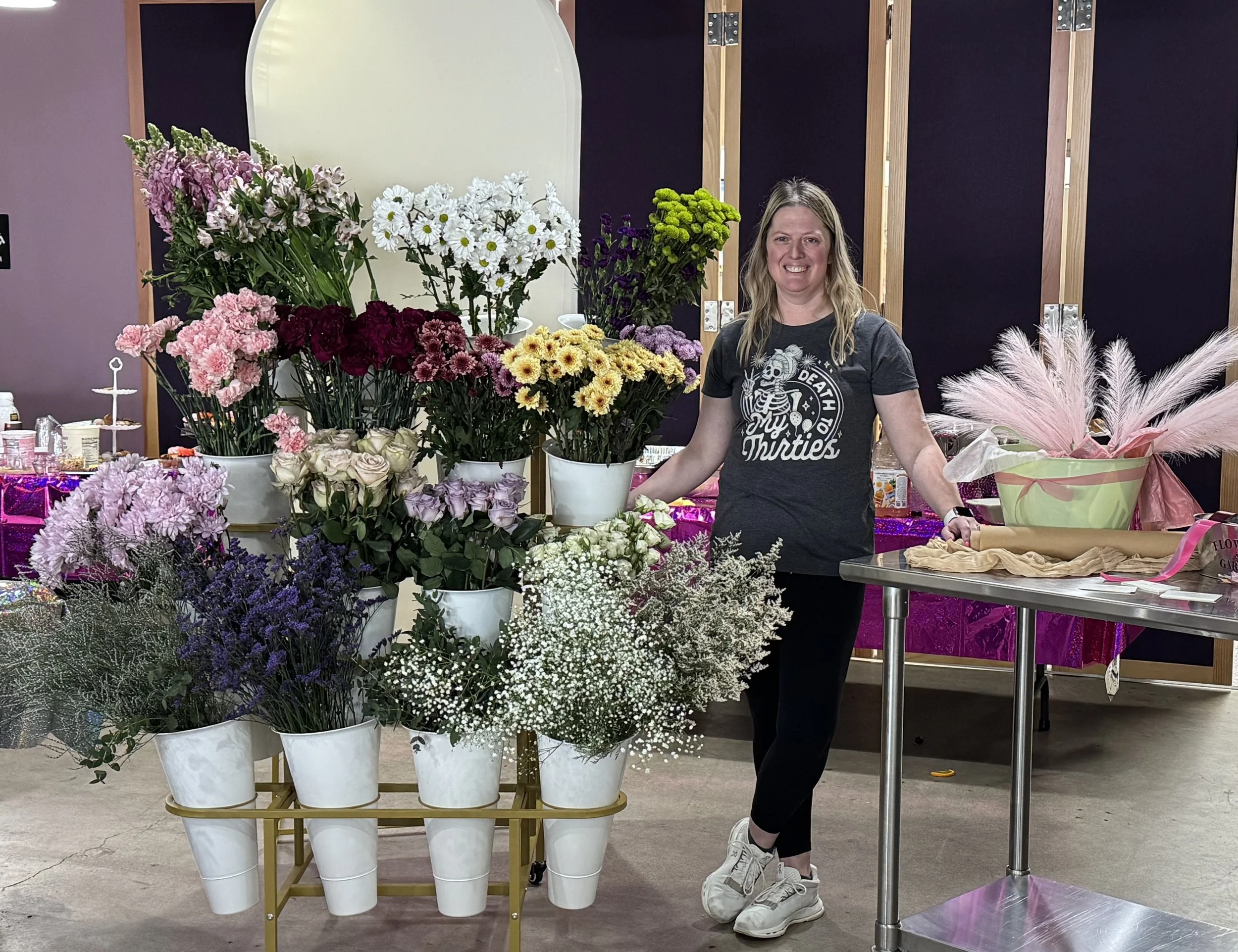 A woman standing next to a display of various colorful flowers in white pots, with a table on the right holding a large green basket with pink pampas grass and a wrapped flower arrangement, in an indoor setting with purple and black walls.