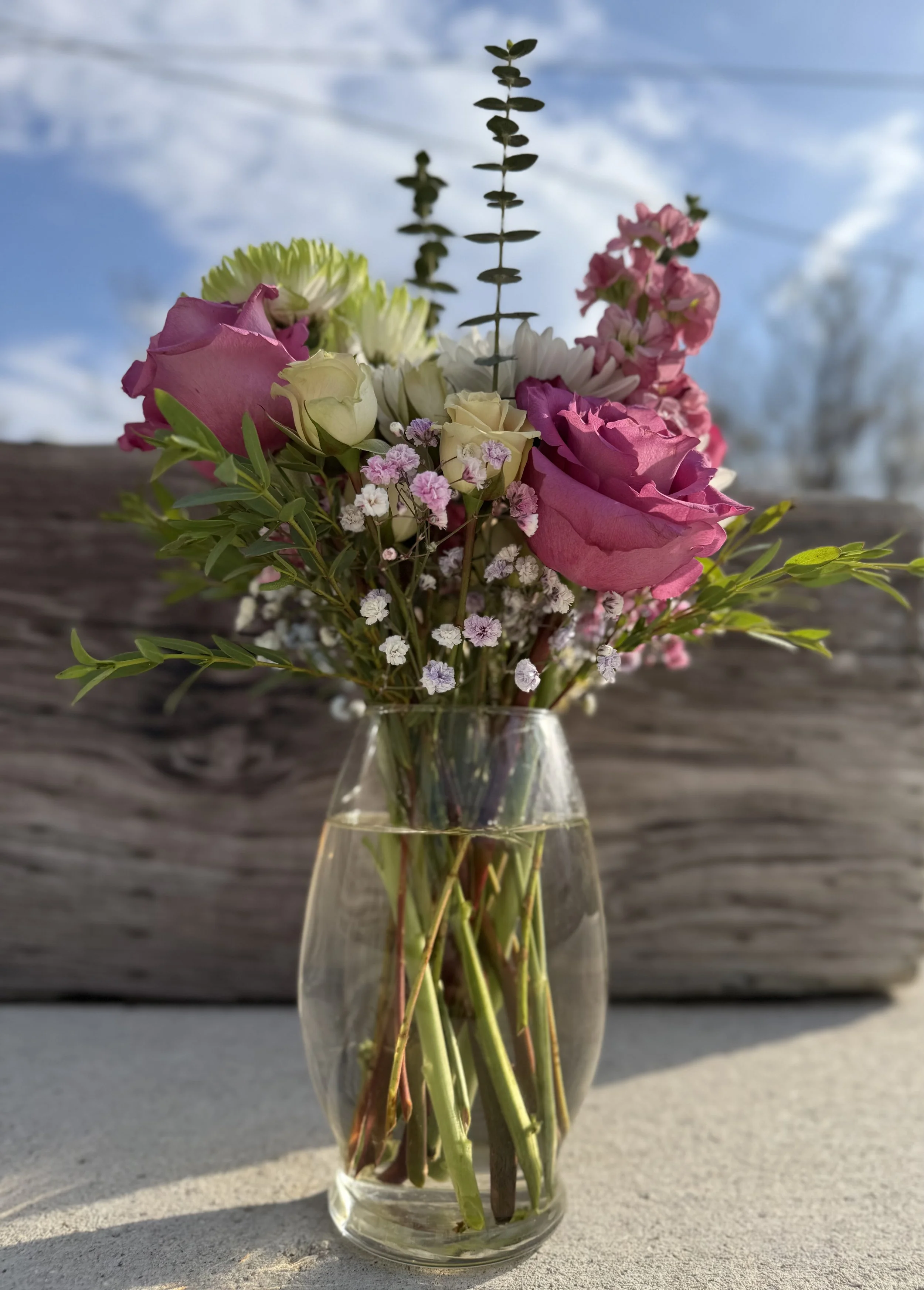 A clear glass vase with a bouquet of pink roses, white roses, small pink and white flowers, and greenery on a surface outdoors, with a weathered wooden background and a partly cloudy sky.