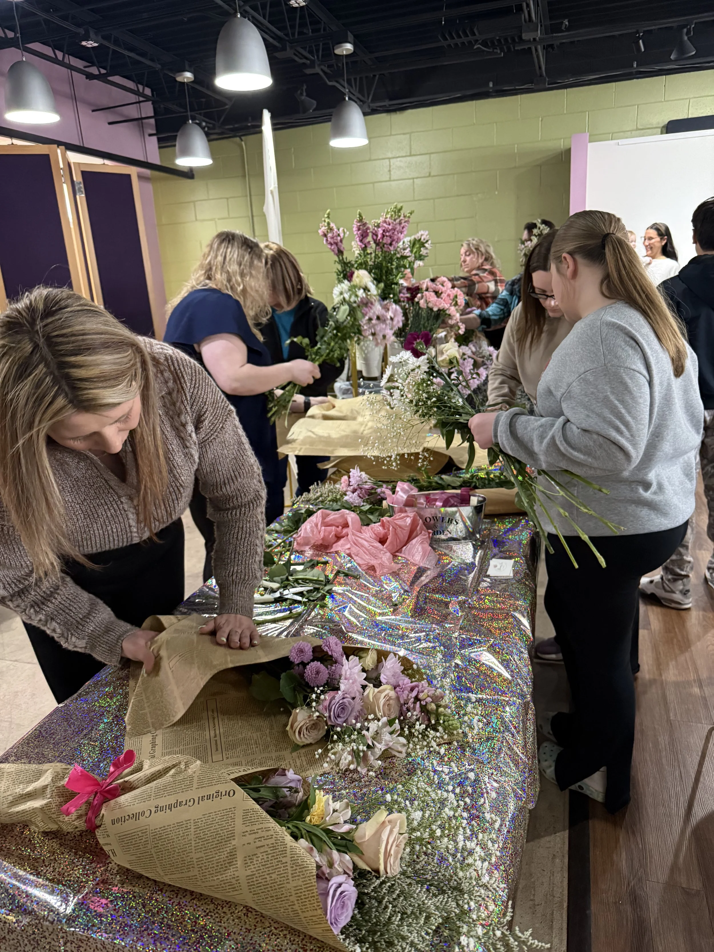People arranging and wrapping flowers on a table covered with a shiny, iridescent tablecloth in a room with green walls and hanging ceiling lights.