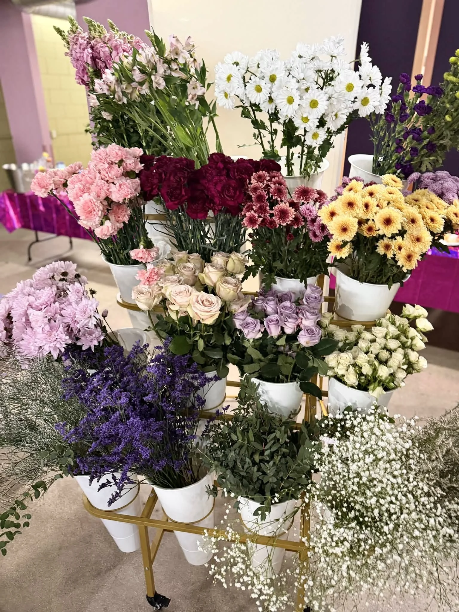 Various colorful flowers in white buckets arranged on a gold-colored, multi-tiered cart, including pink, purple, red, yellow, and white blooms.