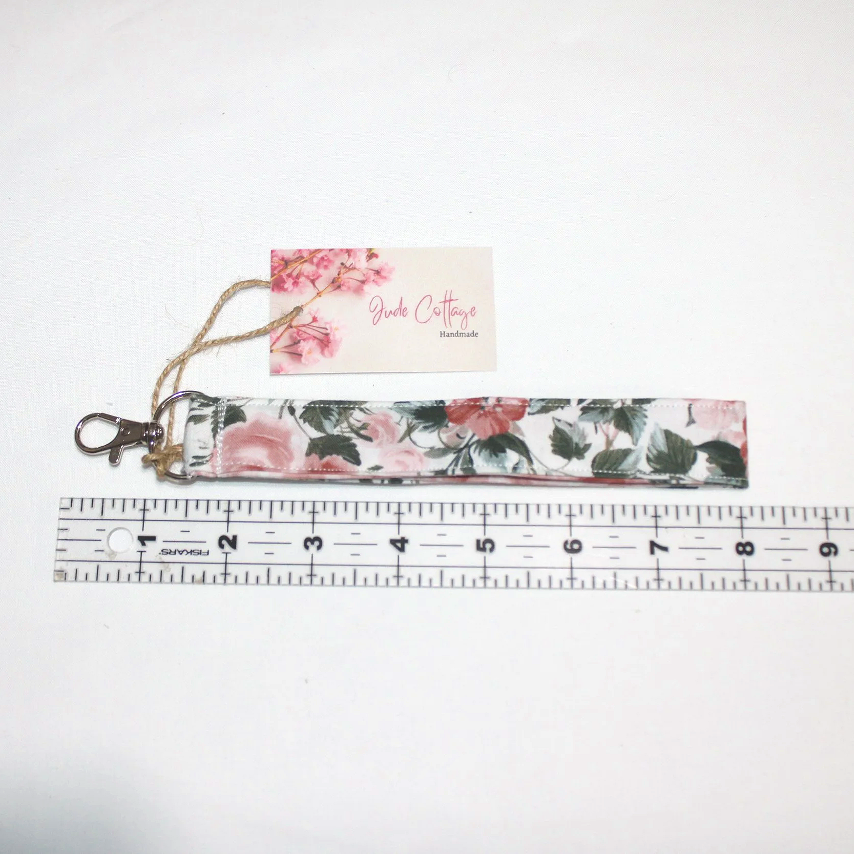 Rose Garden Wristlet made from pink and red rose‑print fabric with an approximately 7.5‑inch strap, metal clasp, and branded tag, displayed on a neutral background shown with a ruler for size.