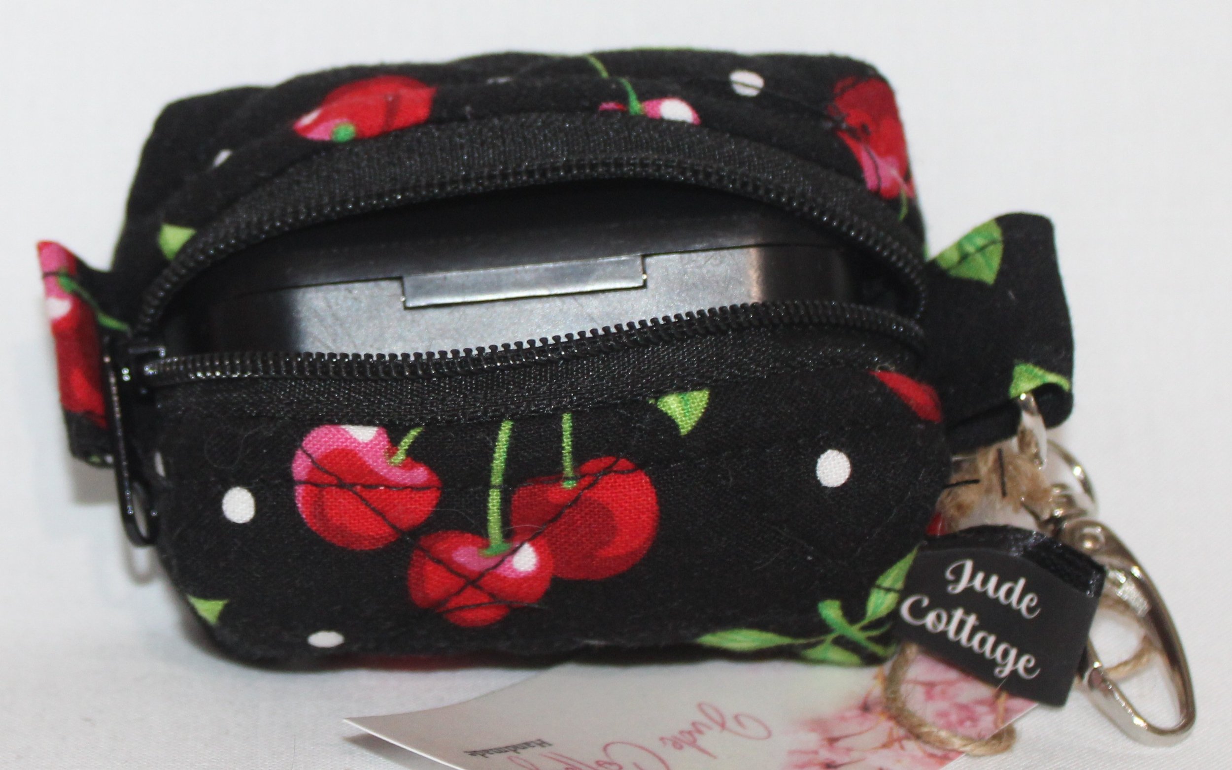 Top open view of the Scattered Cherries Bag Charm showing cherry print on black background with a set of J Lab earbuds inserted for sizing reference.