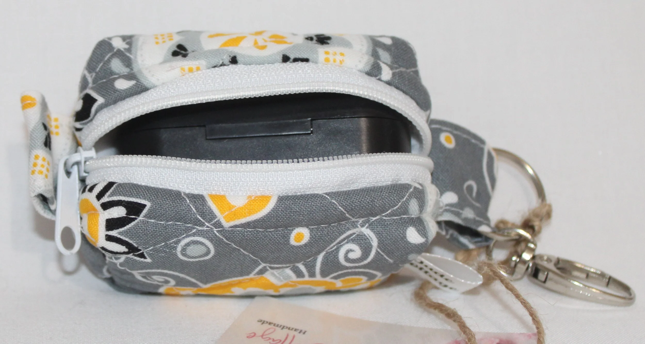 Top open view of the Medallion Bag charm featuring gray fabric with black, white and yellow floral print shown with an J Lab earbud case inserted for sizing reference.