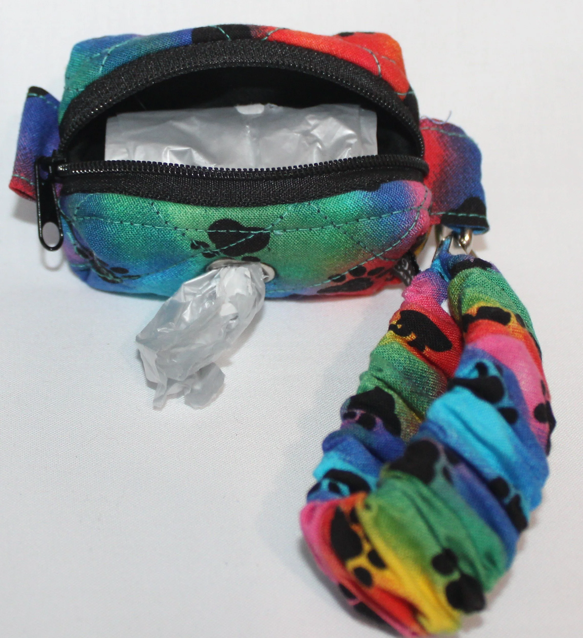 Open view of the Pawsitivity mini boxy pouch in rainbow paw‑print fabric, showing a roll of waste bags inside and one bag pulled through the side grommet.