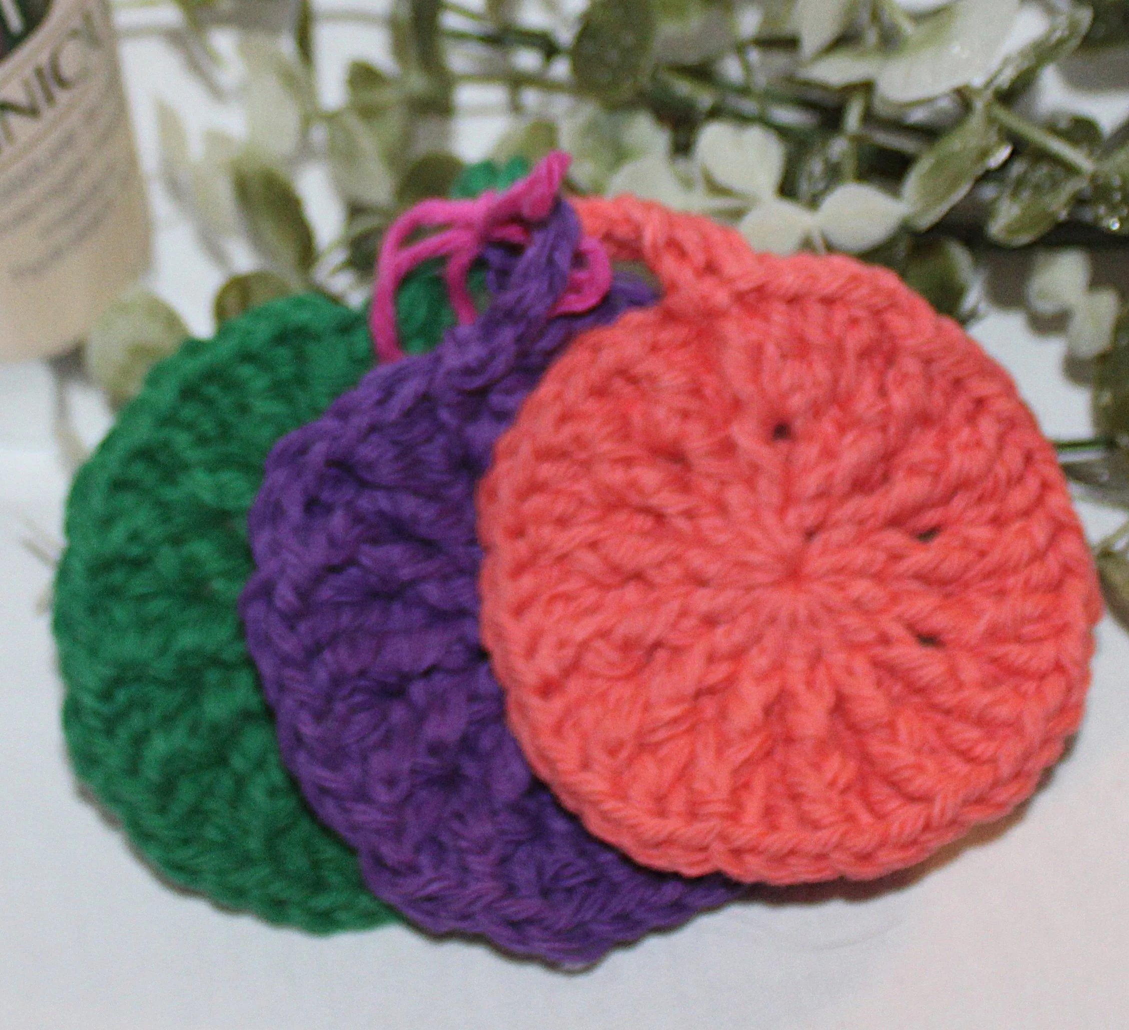 Close‑up of round crocheted assorted colors of green, plum, and coral facial scrubbies with a textured stitch pattern.