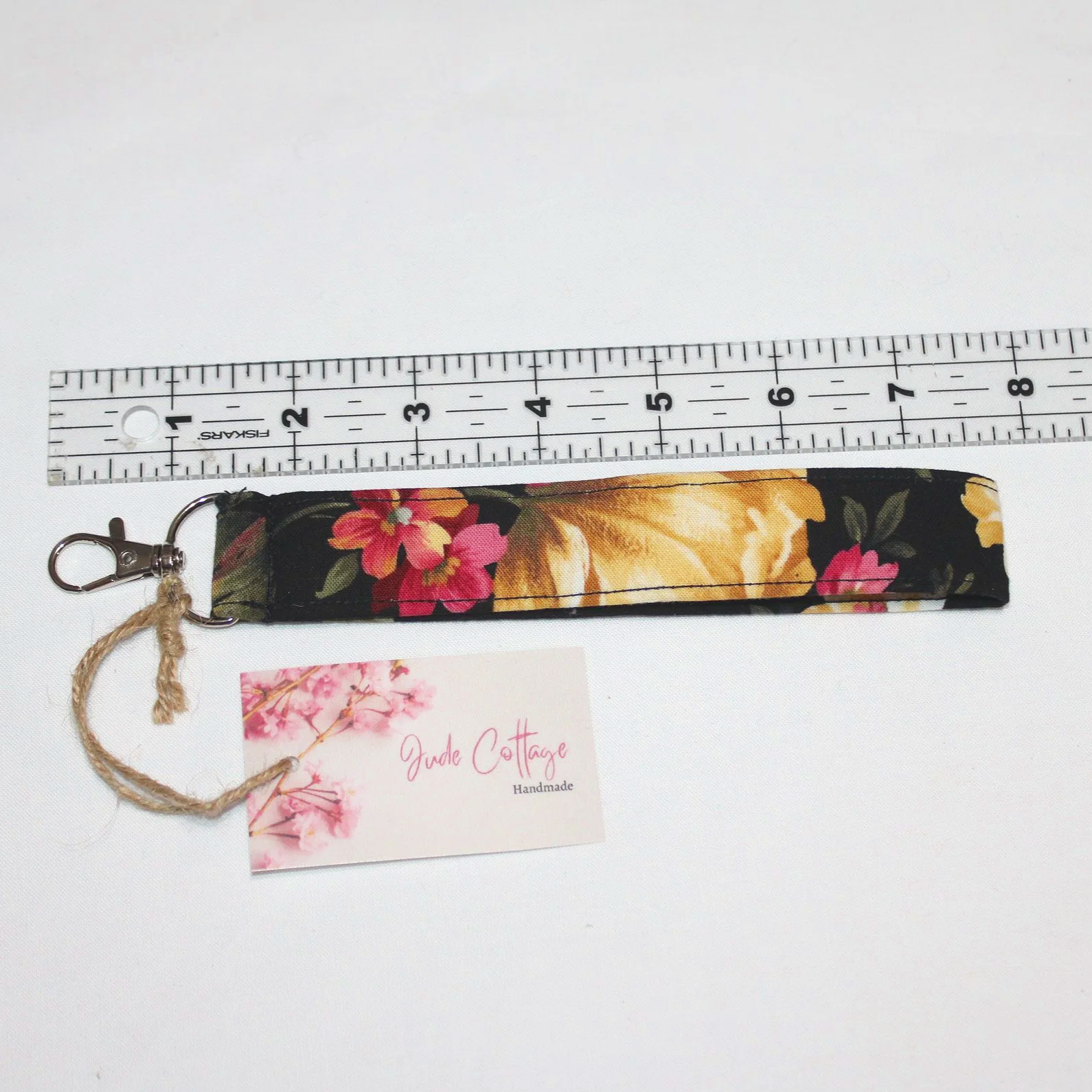 Garden Bloom Wristlet made from colorful floral‑print fabric with an approximately 7.5‑inch strap, metal clasp, and branded tag, displayed on a neutral background shown with ruler to show size.