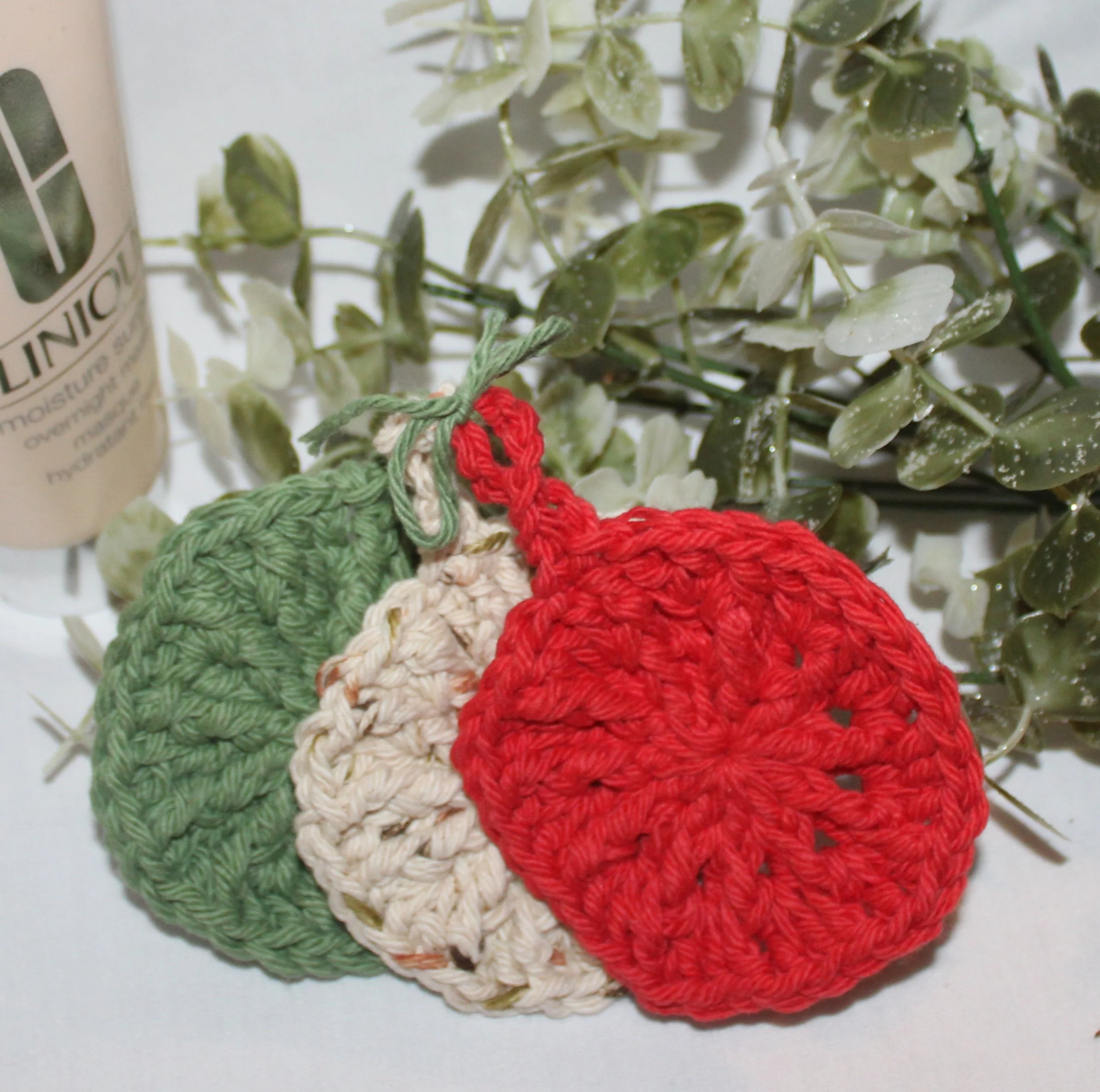 Close‑up of round crocheted assorted colors of red, sage, and sand facial scrubbies with a textured stitch pattern.