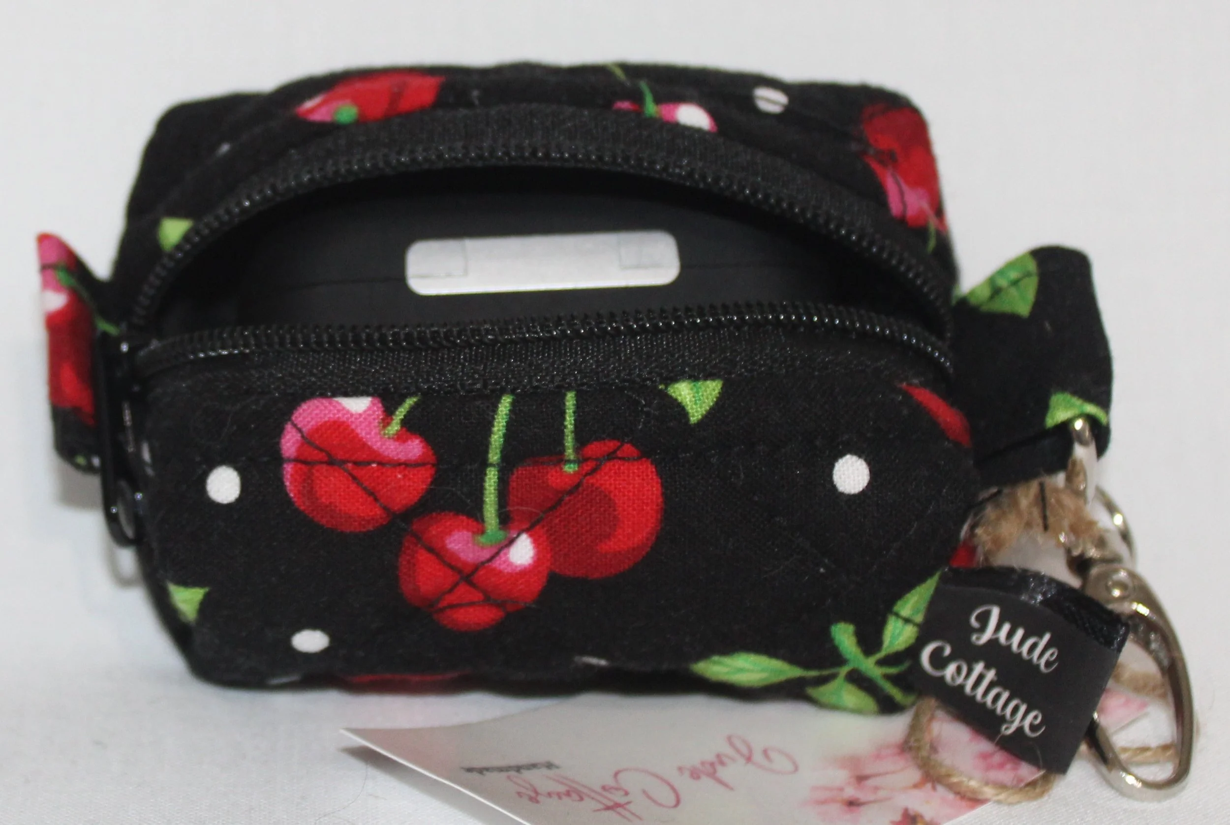 Top open view of the Scattered Cherries Bag Charm showing cherry print on black background with a set of Airpod Pro earbuds inserted for sizing reference.