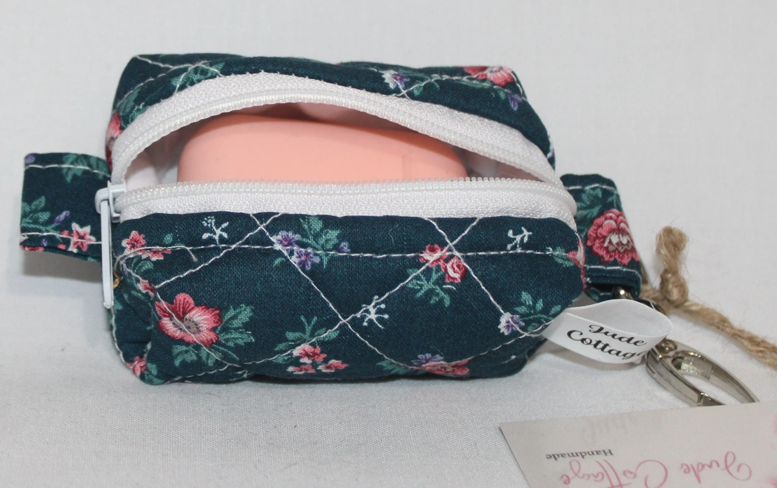 Open top view of the Eliza Bag Charm featuring a dark teal background with pink and lilac floral print shown with an Airpod earbud case inserted demonstrating size.