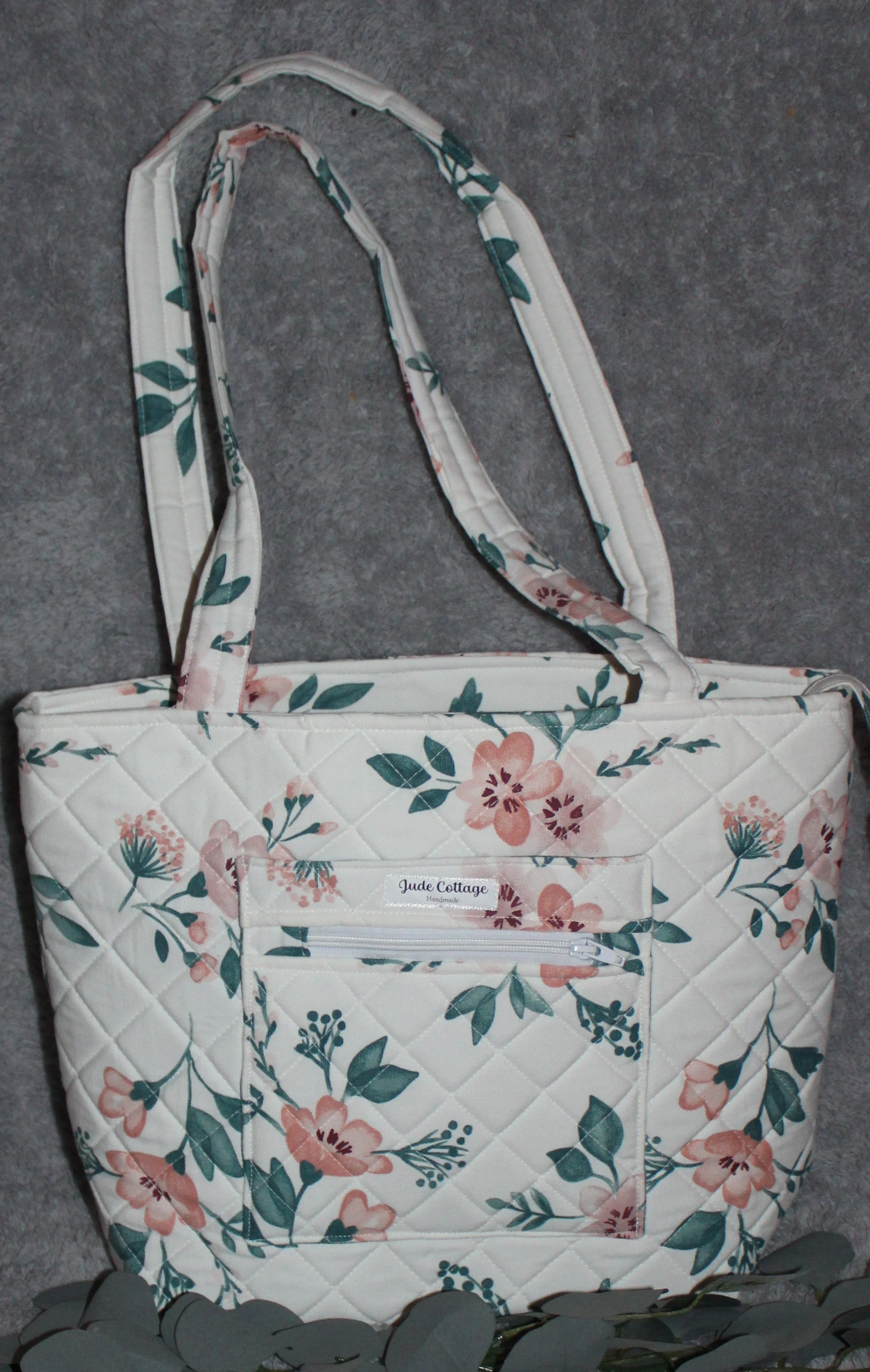 “Photo of the Juliette, a white zippered tote bag with pink floral print, shown standing upright with a top zipper closure, quilted exterior texture, and exterior slip and zippered pockets.”