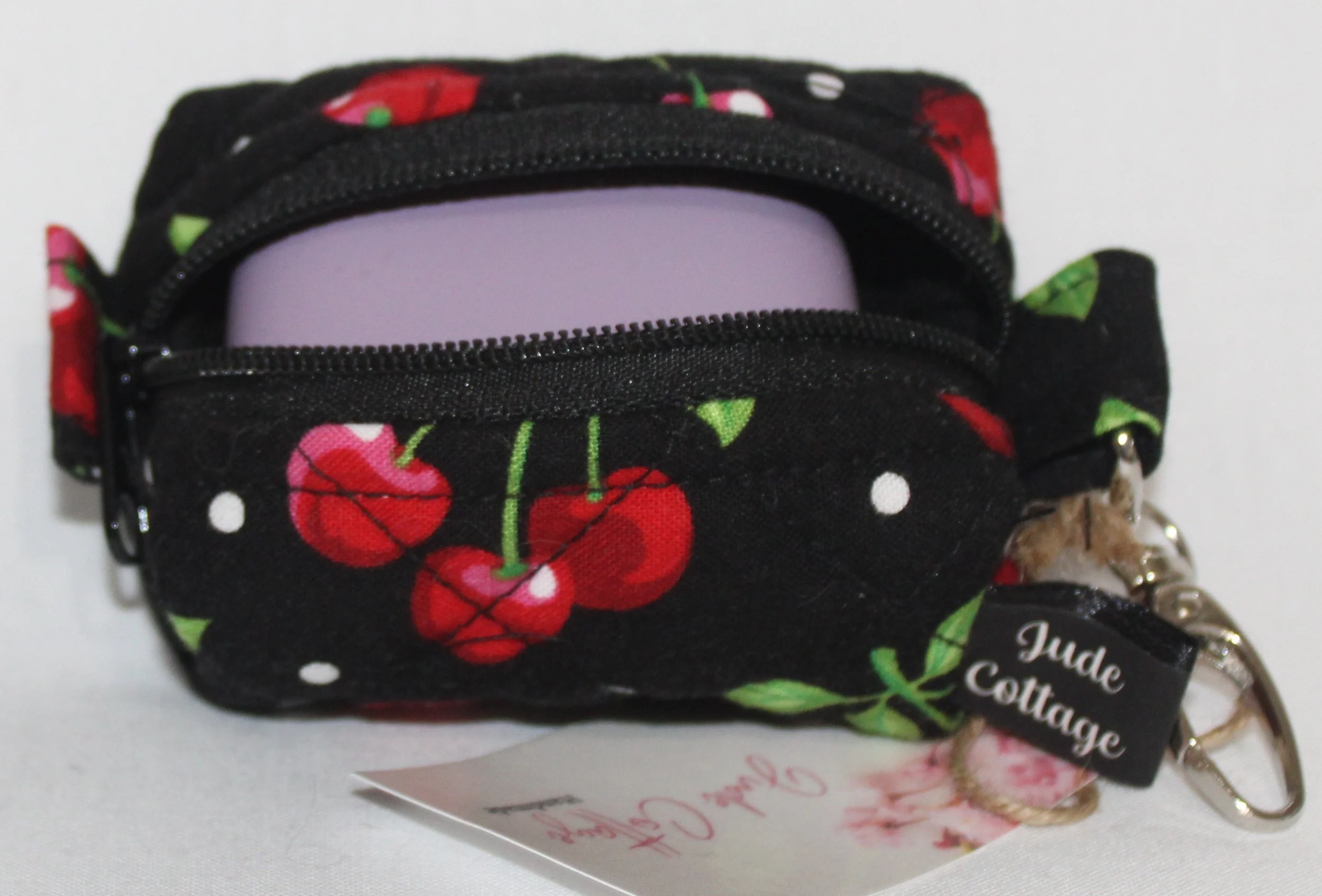 Top open view of the Scattered Cherries Bag Charm showing cherry print on black background with a set of J Lab earbuds inserted for sizing reference.