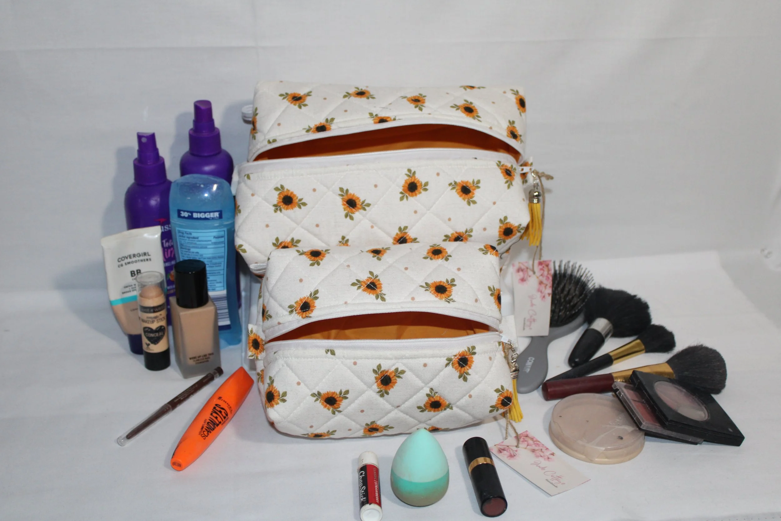 Large and small Sunflower Medley quilted travel pouches in sunflower‑patterned fabric with yellow interiors displayed alongside assorted beauty products.