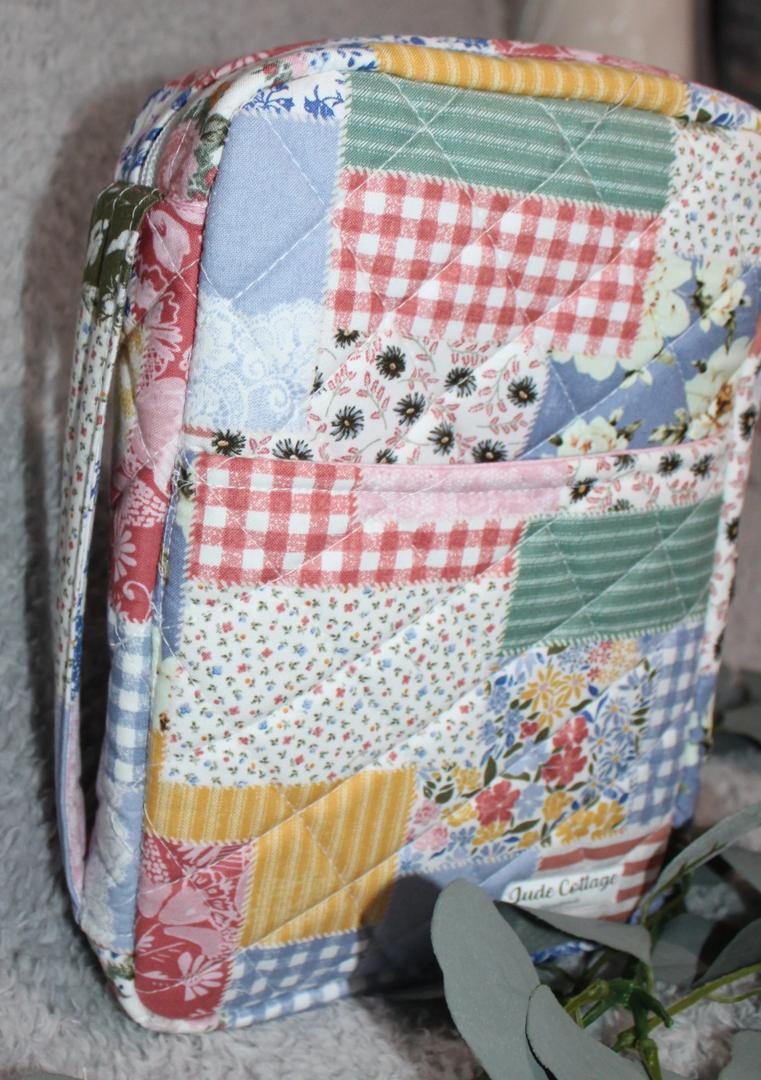 Close-up of a quilted patchwork fabric bag with various floral and gingham patterns and colors including pink, blue, yellow, green, and white, leaning against a textured wall with some greenery in the foreground.