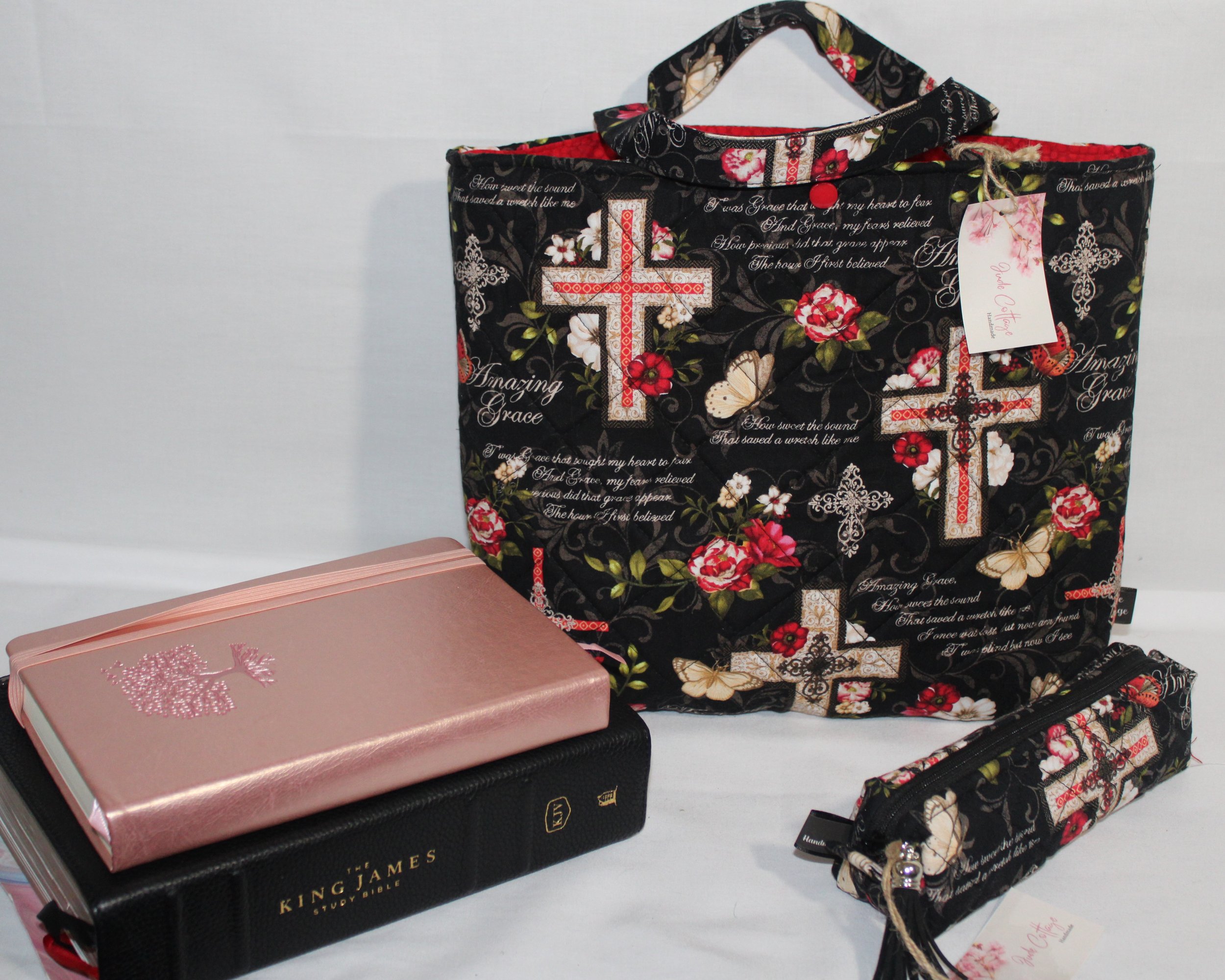 Front view of the Amazing Grace quilted Bible sleeve featuring black fabric with red and white floral prints, cross motifs, and hymn lyrics, shown with a Bible and notebook placed beside it.
