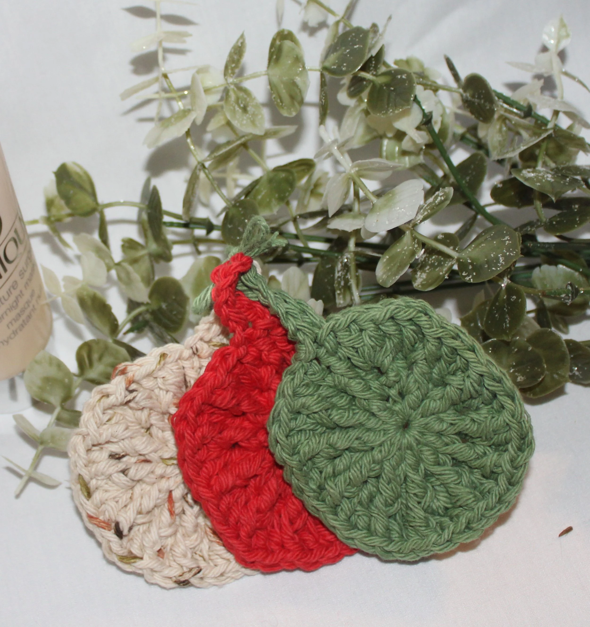 Close‑up of round crocheted assorted colors of red, sage, and sand facial scrubbies with a textured stitch pattern.
