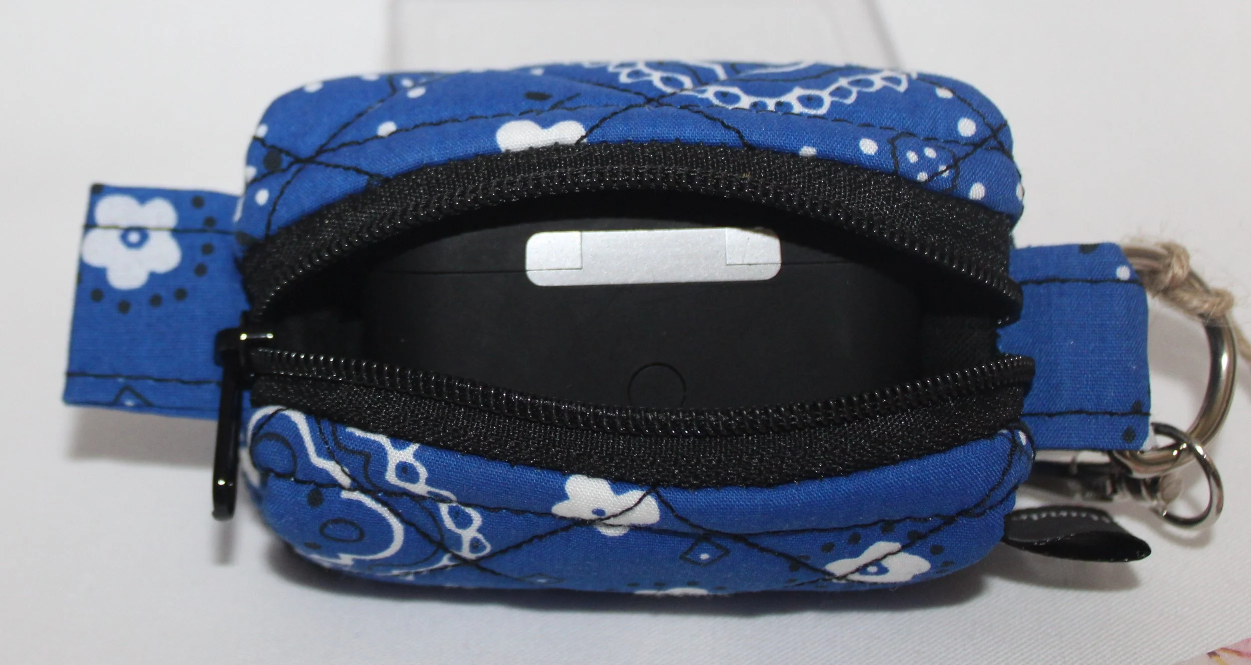 Top open view of the Blue Bandana Bag Charm showing its blue bandana‑print fabric with the zipper fully open. A pair of air pod pro earbuds are placed inside the pouch for size and fit demonstration.