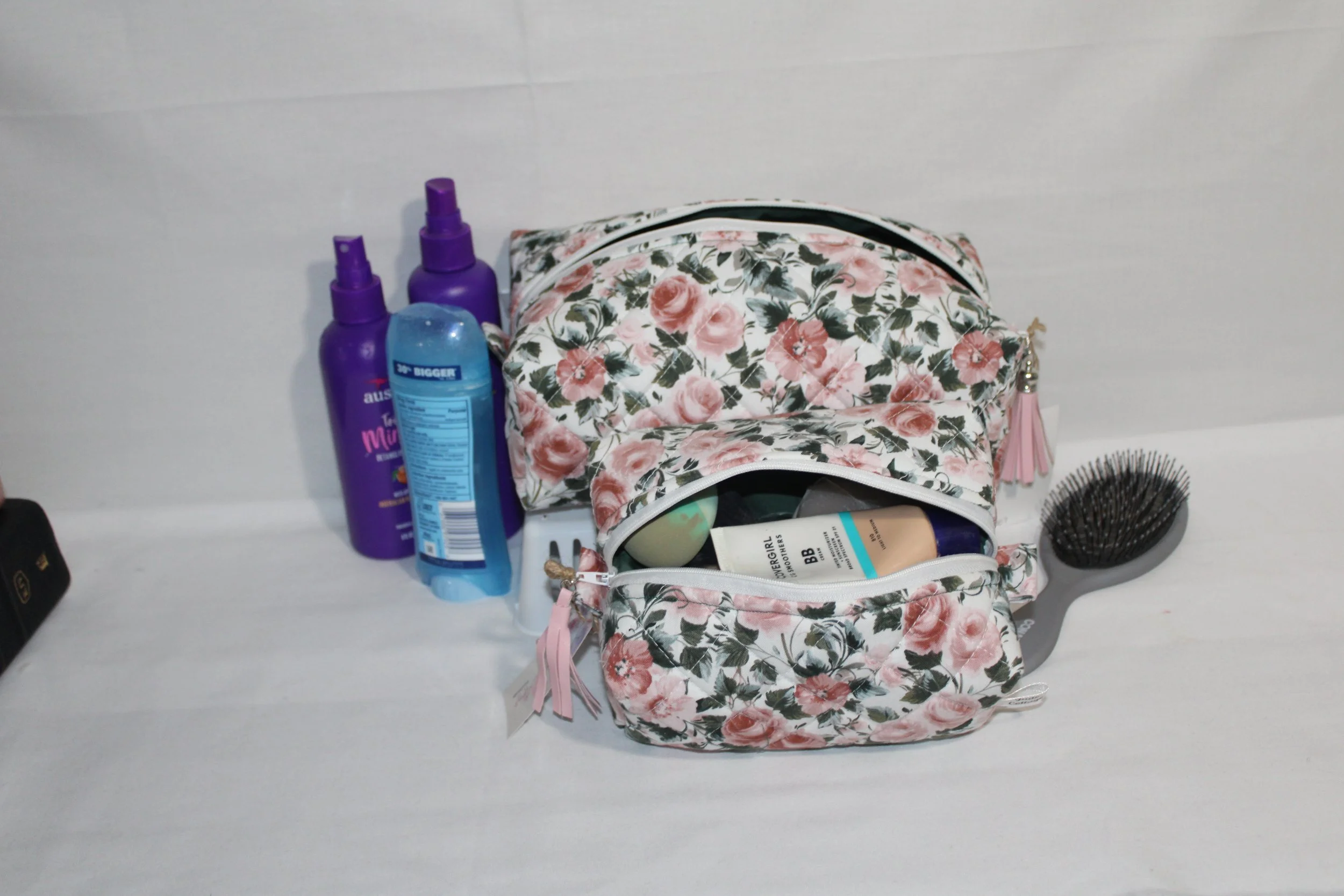 Small Rose Garden quilted travel pouch shown open and filled with cosmetics, displayed beside hair products and a brush on a white surface.