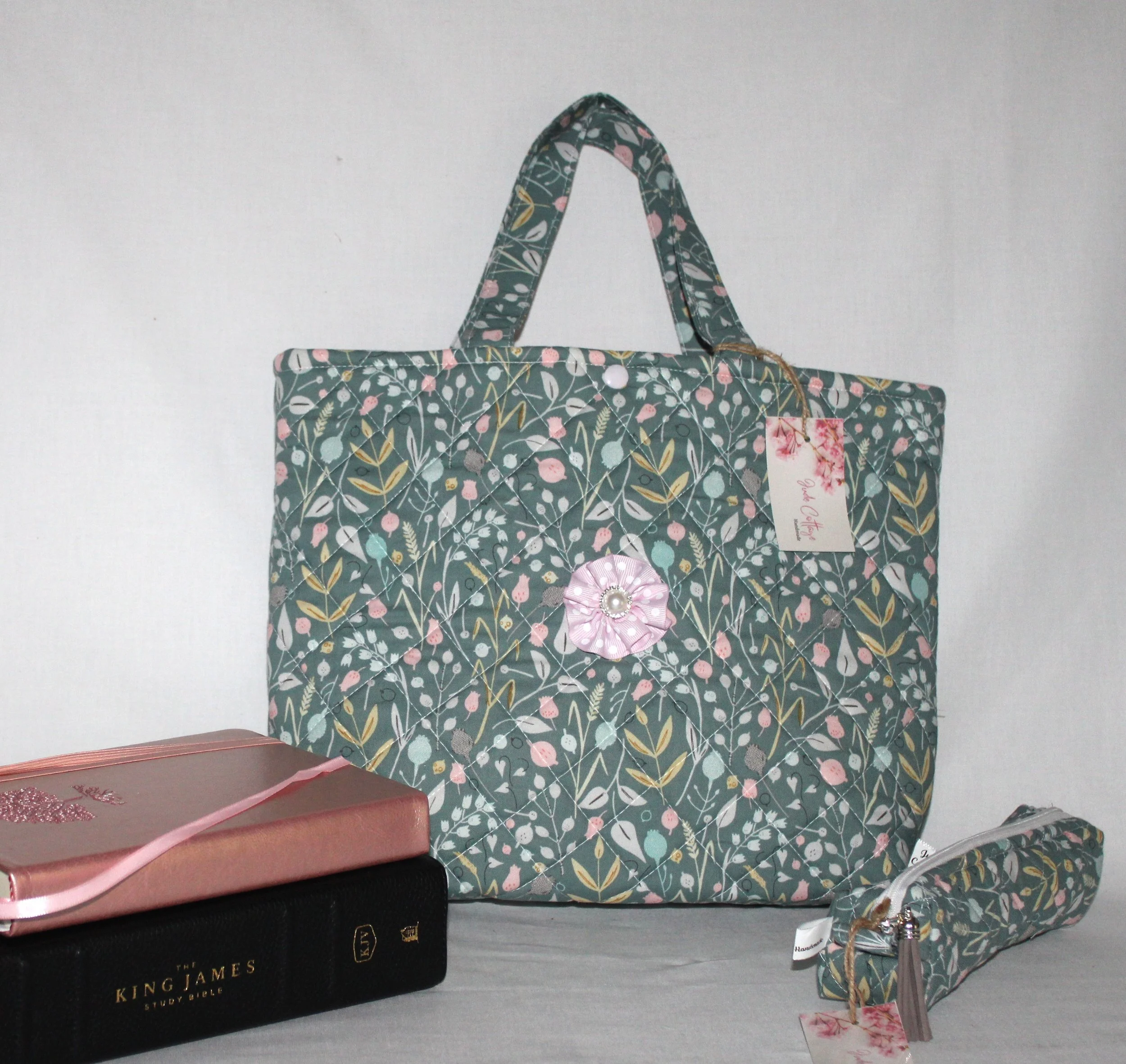Front view of the Meadow Breeze quilted Bible sleeve featuring green floral fabric and two carry handles, shown with a pencil case, Bible, and notebook placed to the side.