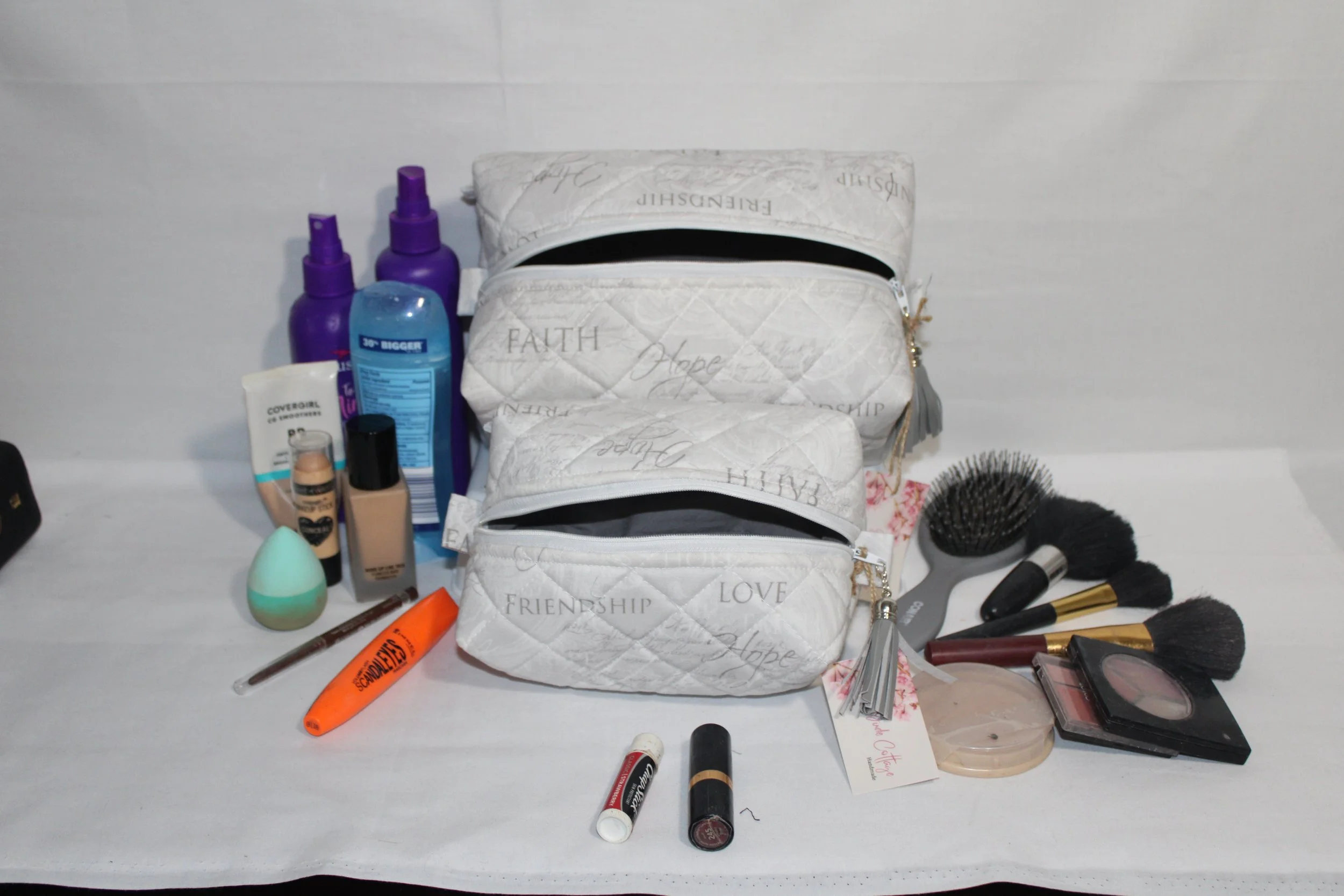Large and small Faith Hope Love quilted cases in light gray inspirational print displayed with cosmetics arranged around them to show size.