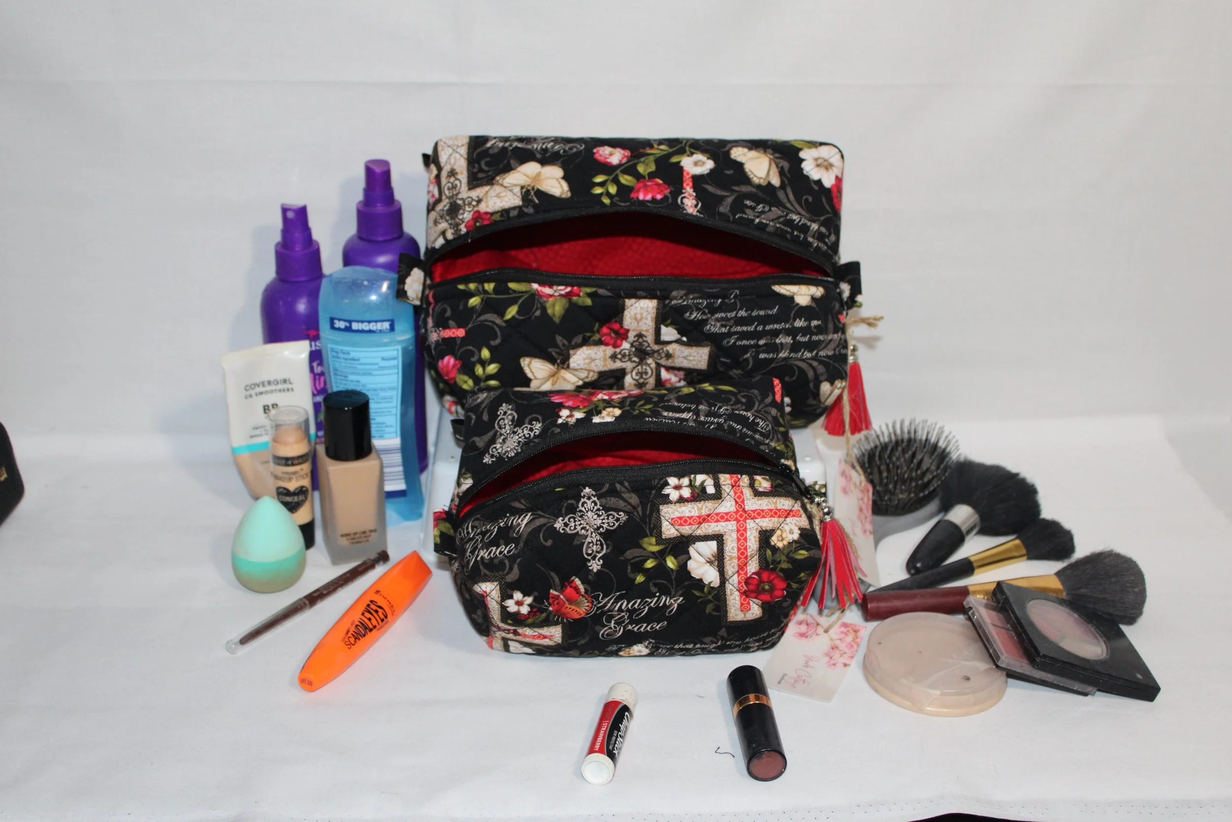 Large and small Amazing Grace quilted travel pouches in multicolor floral print with red interiors displayed alongside assorted beauty products.