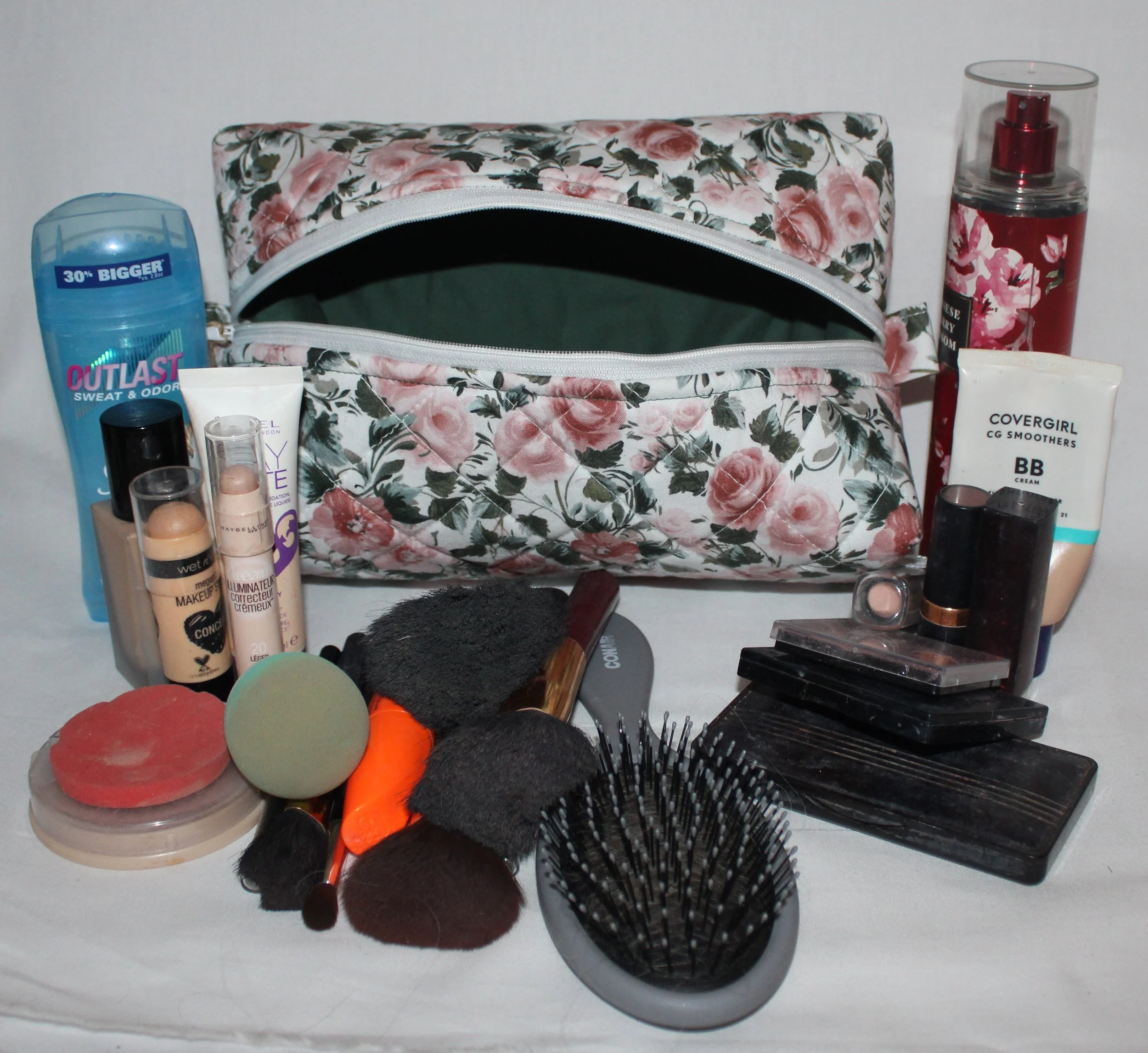 Angled front view of the Rose Garden quilted travel pouch with pink roses and green leaves, shown with a partially open white zipper shown with personal hygiene items to show size.