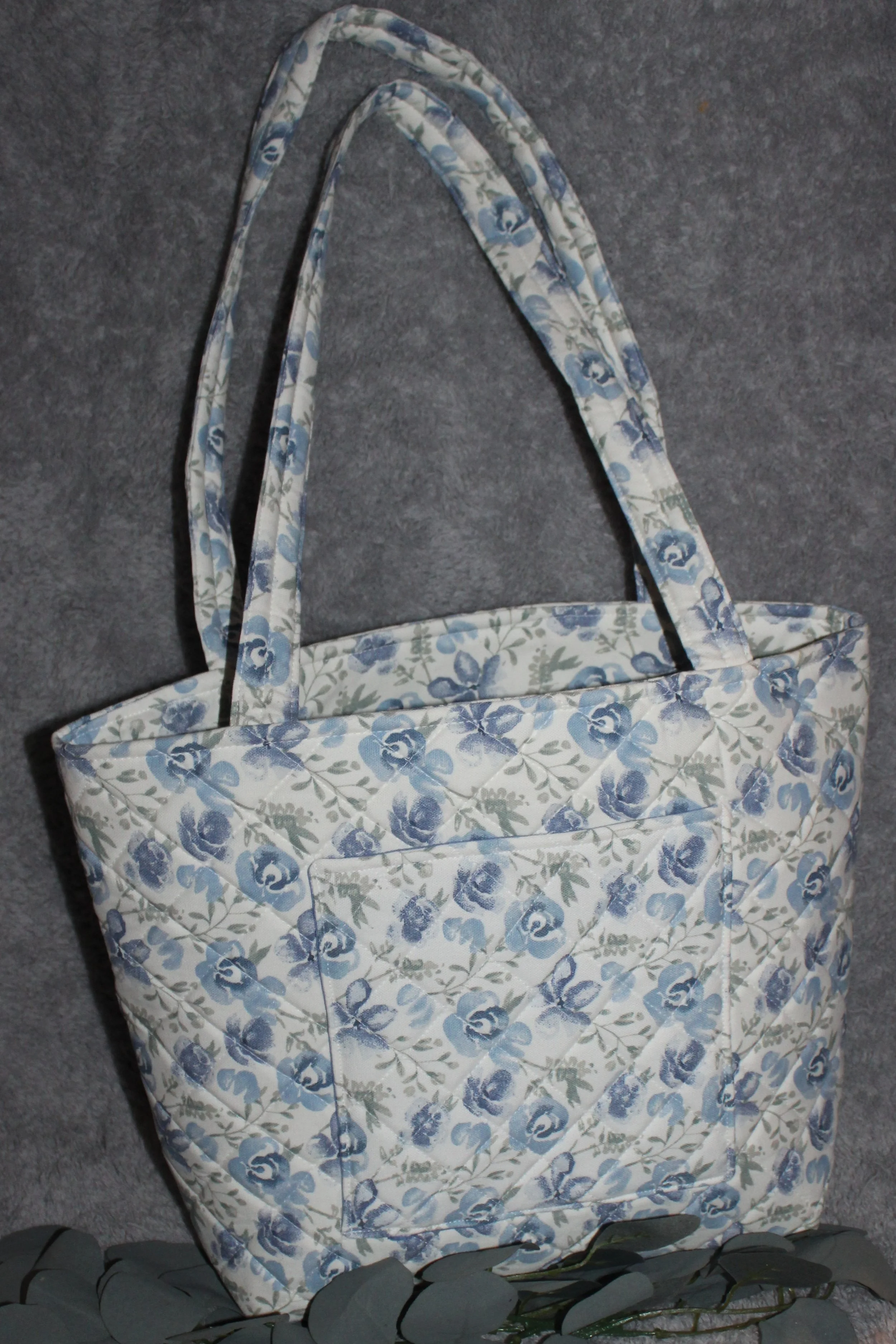 Back view of the Elise quilted tote in white fabric with blue floral pattern, showing the two padded shoulder straps and the exterior slip pocket.
