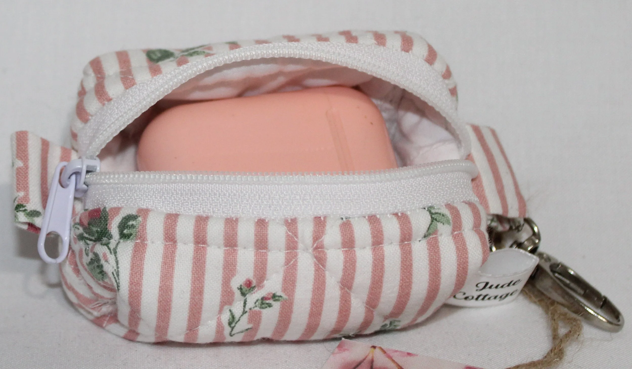 Open top view of the Victoria bag charm featuring pink and white stripes with pink rose bud printed fabric shown with a set of Airpod earbuds inserted for sizing reference.