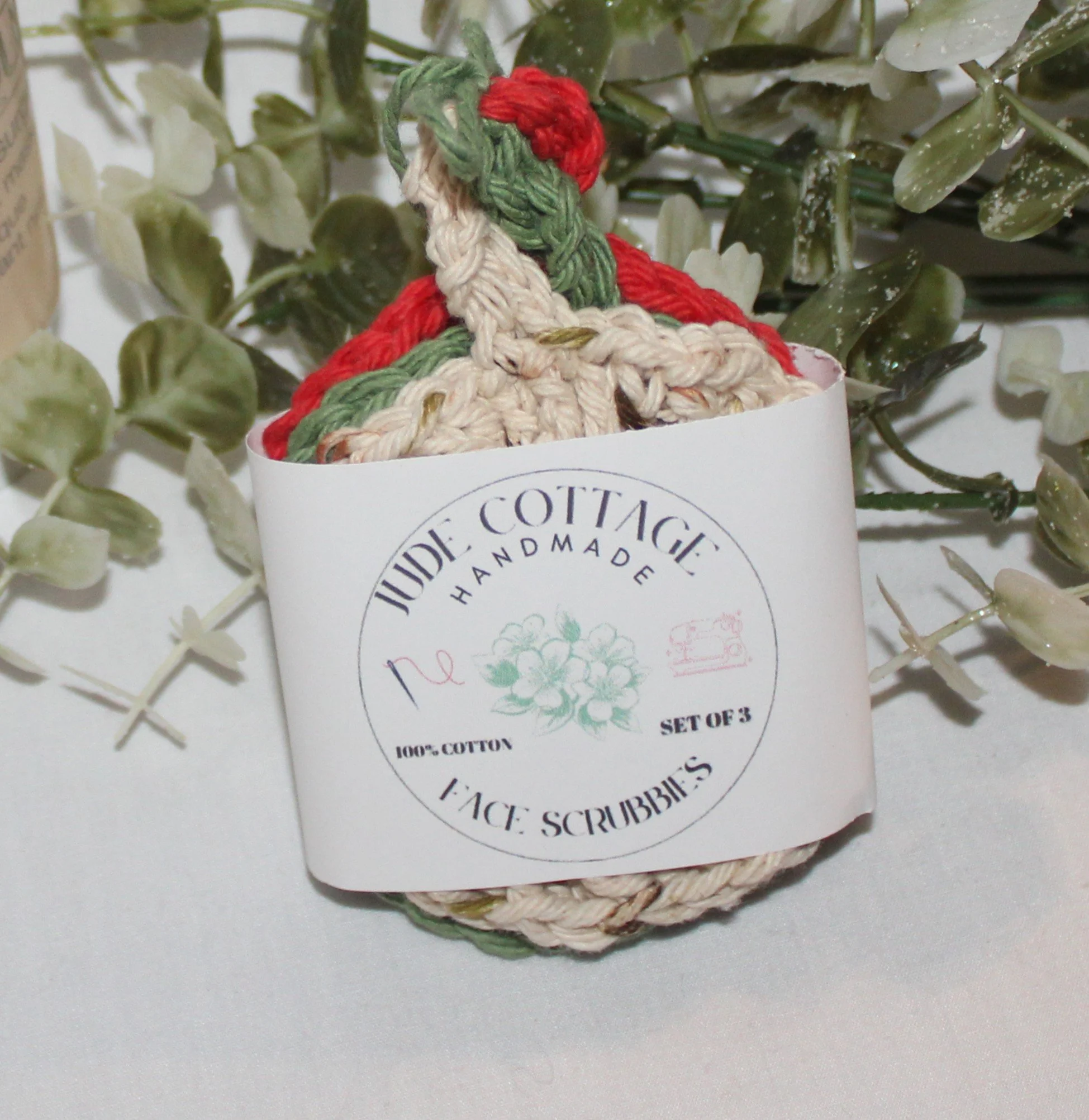 Close‑up of round crocheted assorted colors of red, sage, and sand facial scrubbies with a textured stitch pattern shown with label.